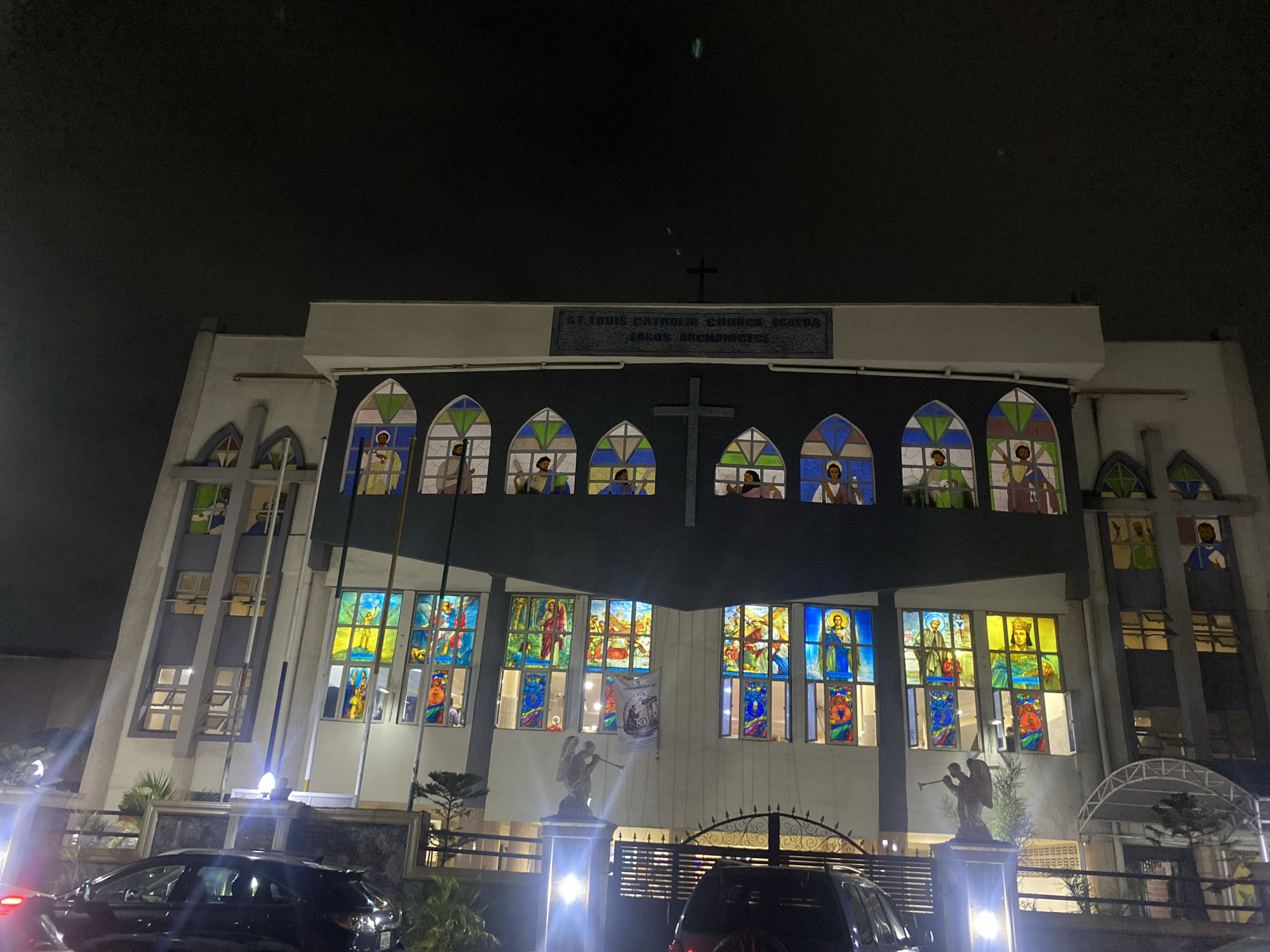 The image depicts the front facade of St. Louis Catholic Church in Ikoyi, Lagos, Nigeria at night. The building is illuminated, highlighting the stained-glass windows depicting religious figures and scenes. A large cross is visible on the upper part of the building, above the windows. A stone plaque identifies the church name and location. The church has a symmetrical design. At ground level, there are cars parked in front of the building. There is an angel statue on the left of the gate.