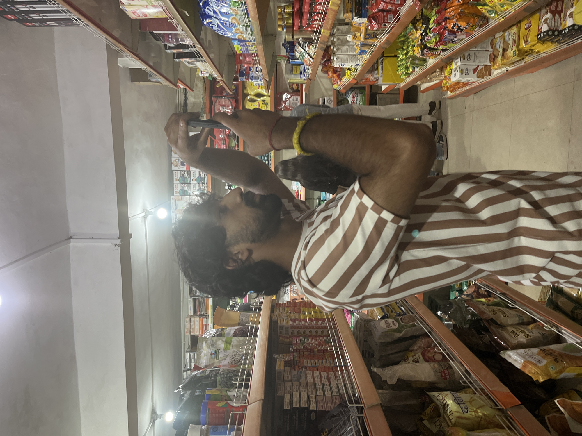 The image shows a person in a supermarket, holding a phone and taking a picture. The person is wearing a brown and white striped shirt and has dark, curly hair. They are standing in an aisle of the supermarket, with shelves filled with various packaged goods on either side. The shelves are stocked with a variety of items, including snacks, cereals, and other packaged foods. The lighting in the supermarket appears to be artificial, consistent with an indoor setting. The overall scene suggests a casual shopping experience, possibly a moment of documenting items of interest or making a purchase. There are no clear indicators of the time of day or weather outside.