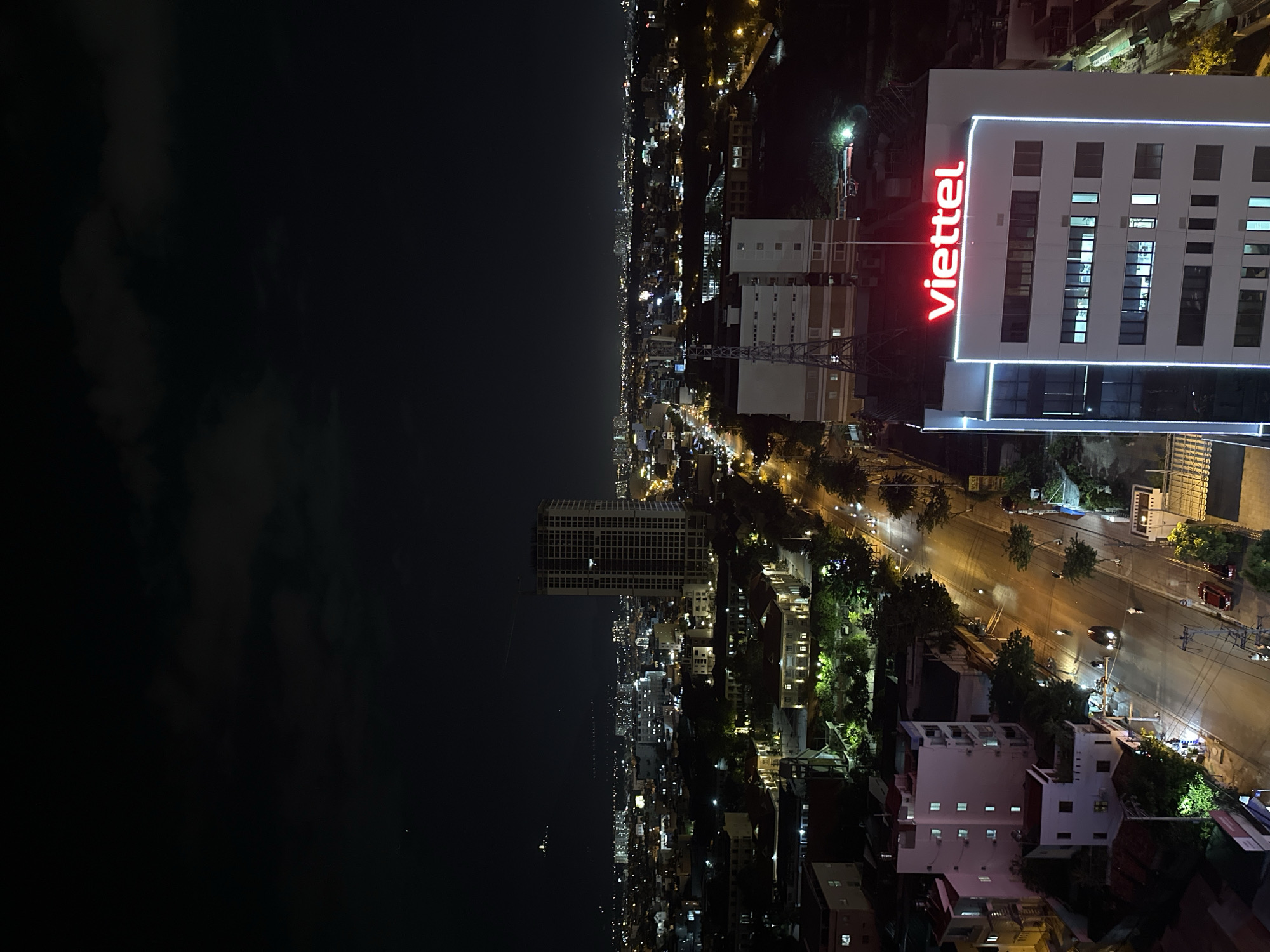 The image is a night-time aerial view of a city. In the foreground, a modern white building with a bright red "viettel" logo is prominently visible. The building has multiple floors with numerous windows, some of which are illuminated. To the left and below, there are residential buildings with red roofs and illuminated windows. A street runs through the scene, with a few vehicles, including motorcycles, visible. In the midground and background, a dense urban landscape with numerous buildings and streetlights stretches towards the horizon, all under a dark night sky with scattered clouds. The overall impression is of a busy, illuminated city at night.