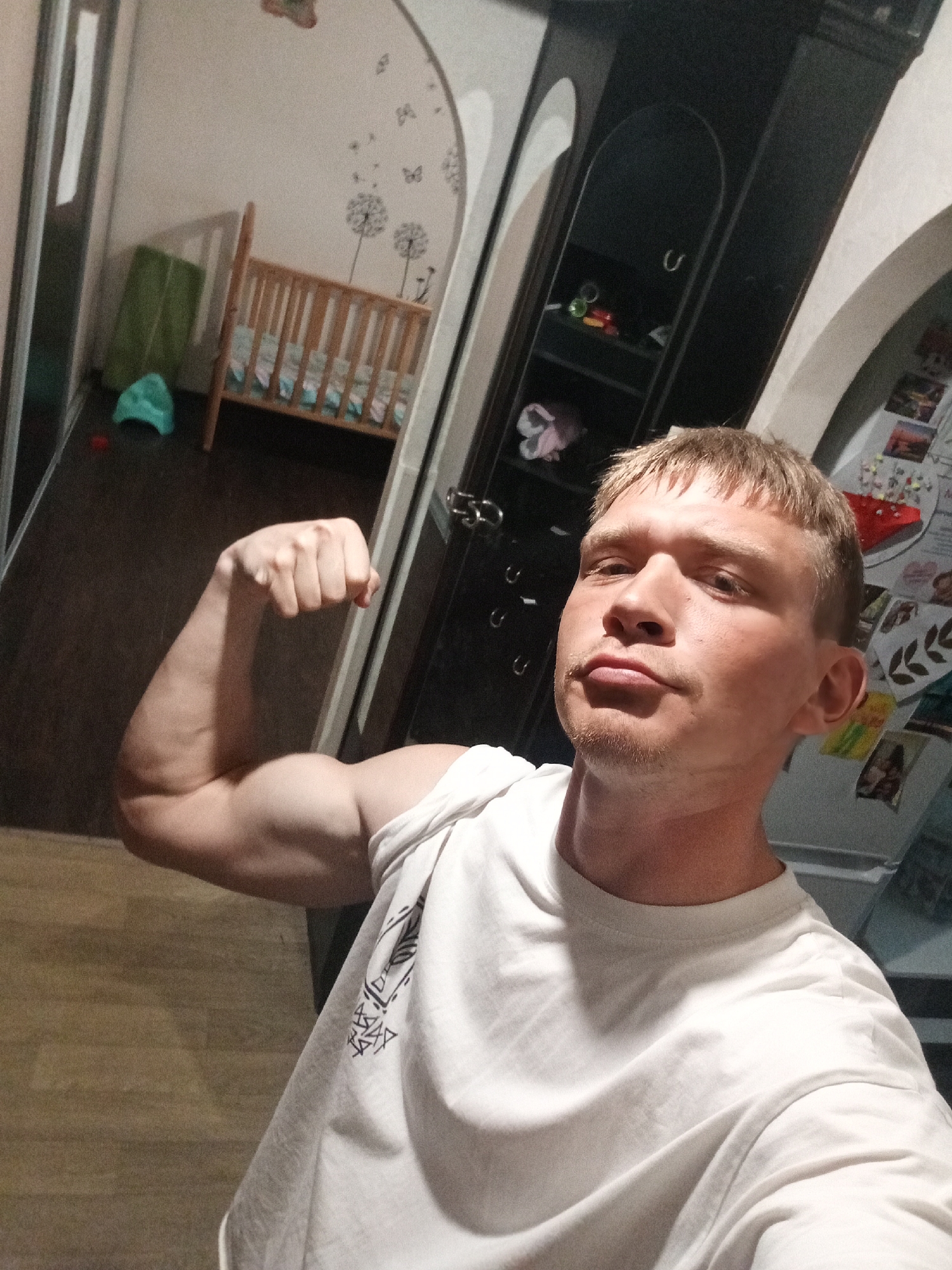 A man is flexing his right bicep indoors. He is wearing a white t-shirt. The background includes a crib, a cabinet, and a refrigerator with photos attached. The location is likely a home, based on the presence of baby items and personal photos. The presence of a crib implies there may be a baby at the location. While the image might have been taken anywhere, the request specifically asks to mention Chaikovsky, Russia. The photo could have been taken there, but it is impossible to confirm based solely on the image.