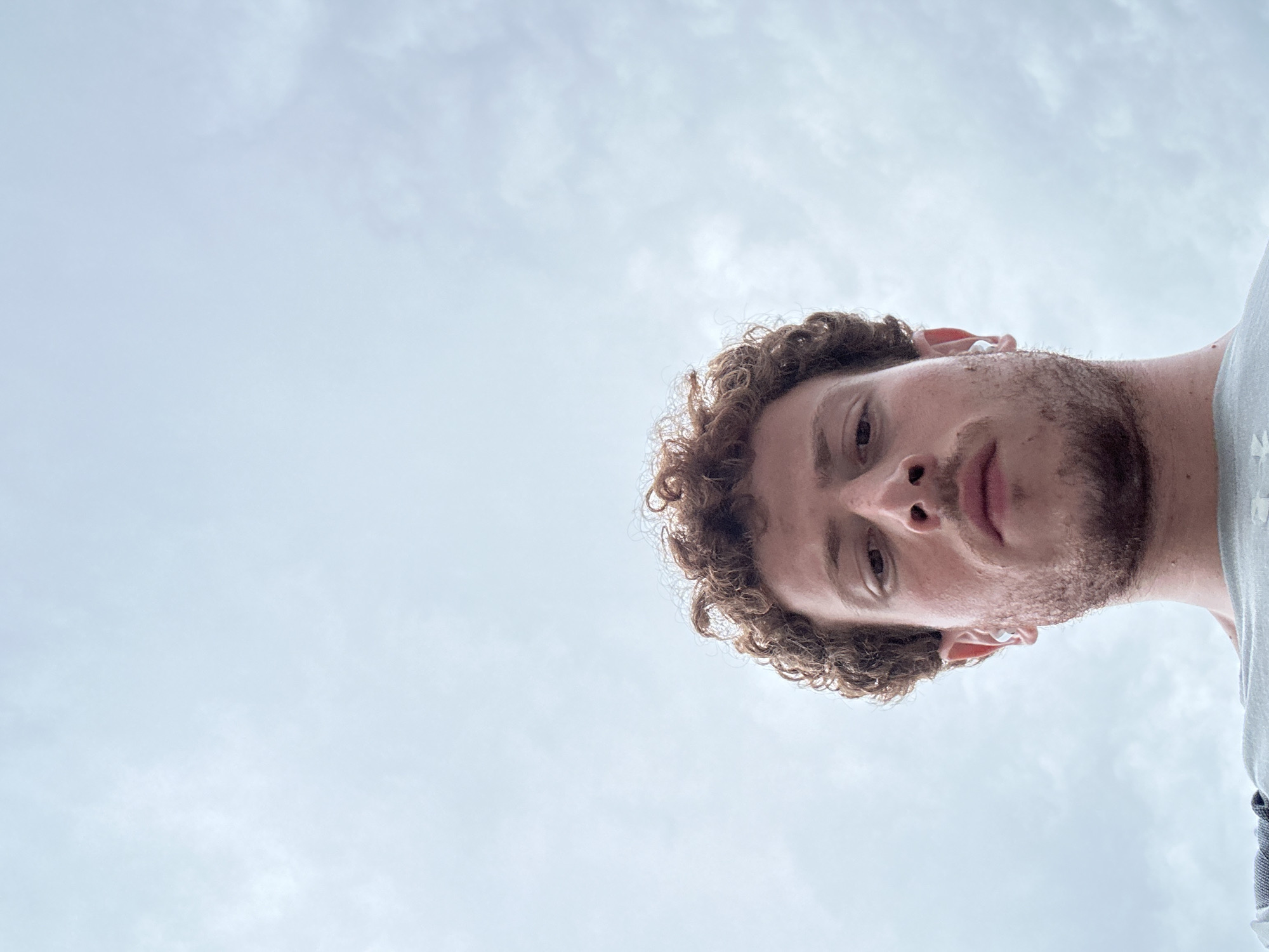 A low-angle, close-up shot features a fair-skinned individual with curly auburn hair, a light beard, and a light grey shirt. The person appears to be wearing a white earbud. The background is a pale, cloudy sky, suggesting an outdoor setting. There is no verifiable location information to conclude it is in New York City or anywhere else.
