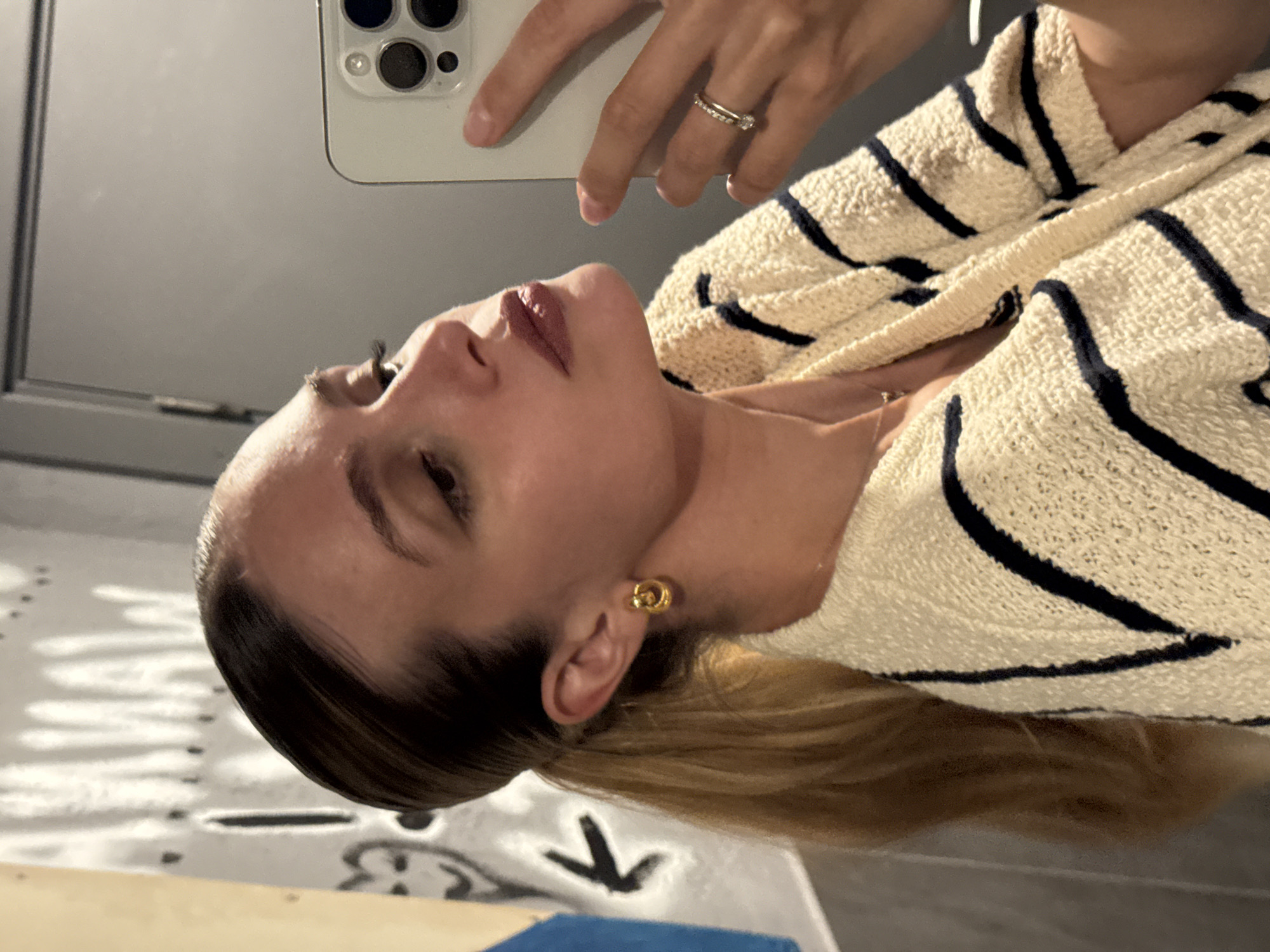 A woman with blond hair and fair skin is taking a selfie in what appears to be an interior space with a door, possibly a bathroom. She is wearing a striped beige sweater and gold hoop earrings. She is holding a cell phone in her left hand and her face is angled upwards towards the camera. A diamond ring is visible on her left ring finger. The location and city, Tbilisi, Georgia, cannot be determined from the available information.