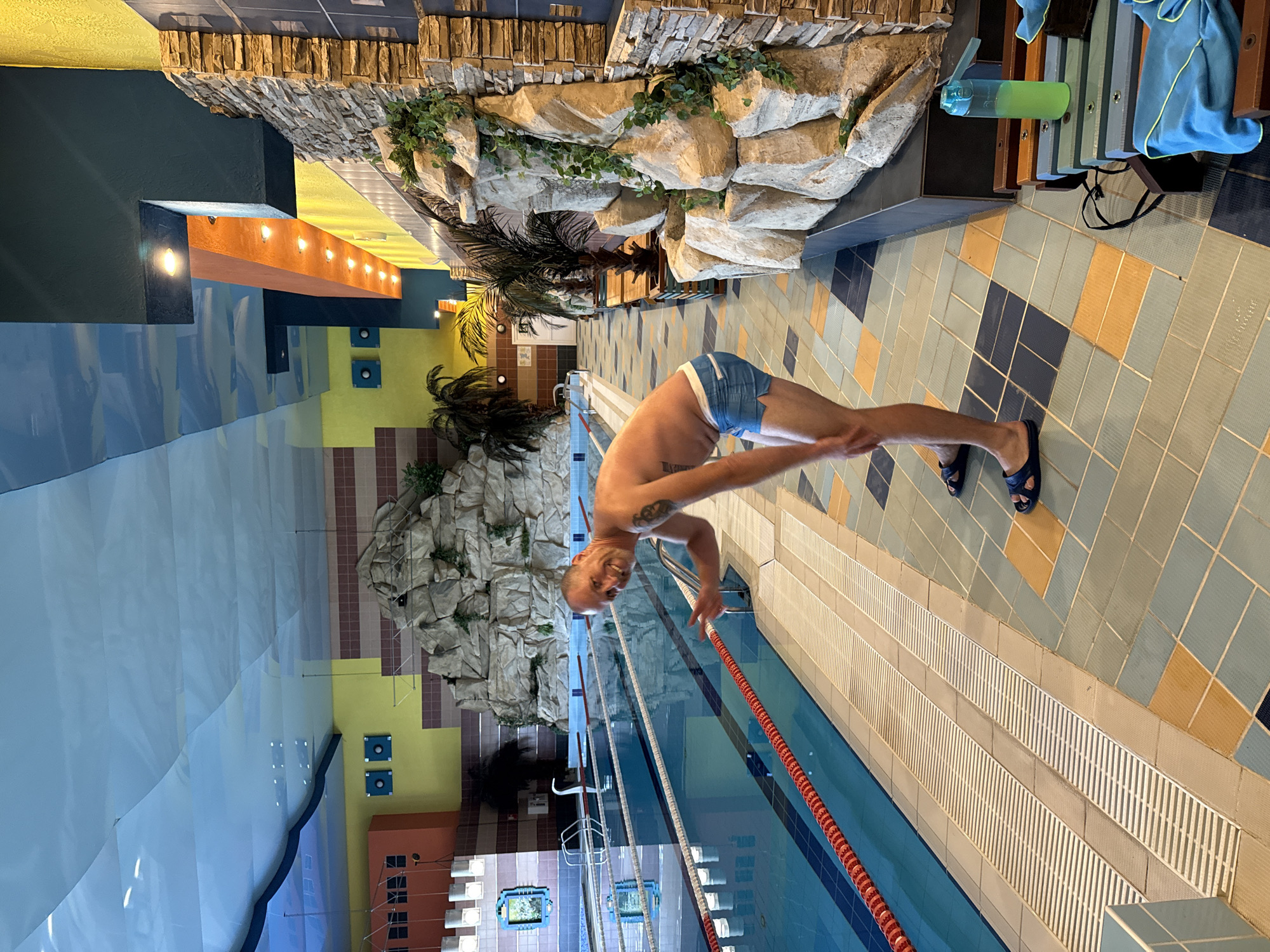 A man is posing in front of a swimming pool, likely indoors due to the artificial rock formations and palm trees. He is wearing blue swim trunks and smiling at the camera, with his left hand raised. The pool lanes are clearly marked, and the water is a vibrant blue. The surrounding area features colorful tiles on the floor and walls, as well as a textured ceiling that mimics a sky or water pattern. The man appears to be alone in the frame, and the overall atmosphere is casual and recreational. There are no visible elements in the image that would suggest a specific location in Chaikovsky, Russia, or any other city for that matter.