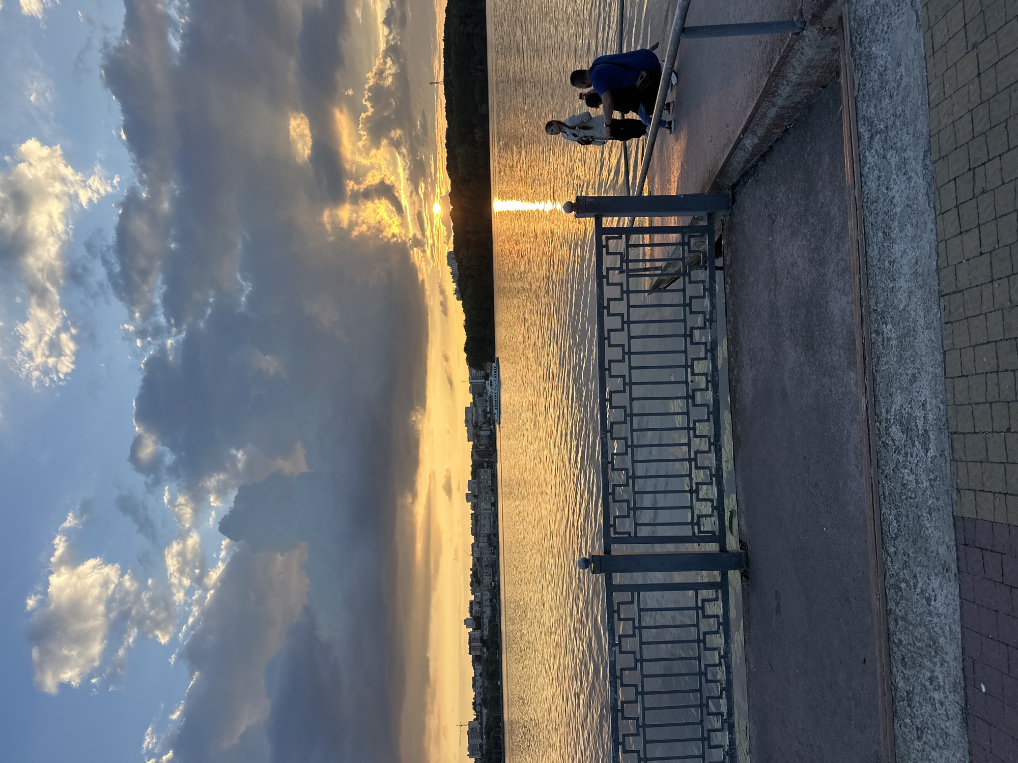 A vertical, medium shot captures a picturesque sunset over Ternopil Lake in Ternopil, Ukraine. The sun, a bright orange orb, is partially obscured by dramatic, dark clouds that stretch across the sky, allowing golden rays of light to pierce through, illuminating the water and the surrounding landscape. A silhouette of a city skyline lines the distant shore.
In the foreground, a grey metal railing borders a concrete walkway. On the walkway, three individuals are present: a person kneeling in a blue shirt, facing away from the camera and towards the lake, seemingly observing the sunset. Beside them, another person, a woman in a light-colored jacket, stands and looks towards the lake. A child in a grey top and black pants stands between the two, also facing the water. The overall atmosphere is serene, with the warm glow of the sunset reflecting on the rippling surface of the lake.