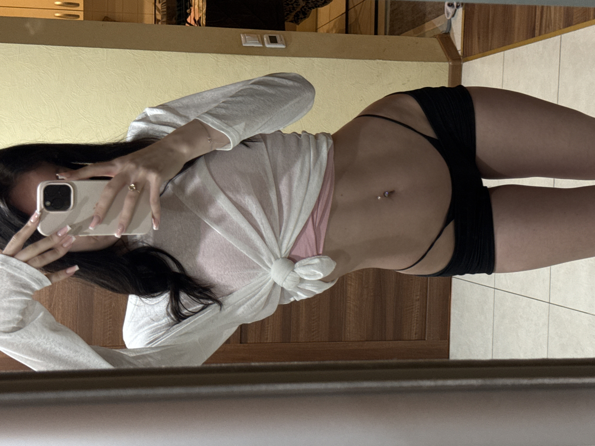 A young woman is taking a mirror selfie in what appears to be an indoor residential setting. She is wearing a white long-sleeved shirt tied at the waist over a pink top, black shorts, and a black thong. She has long dark hair, a navel piercing, and is wearing a ring and a bracelet. The background includes a wooden door, a yellow wall with light switches, and a tiled floor. The image is cropped to focus on the woman's upper body and lower torso. There is no information in the image that would suggest a specific location in the city of Khimki, Russia.