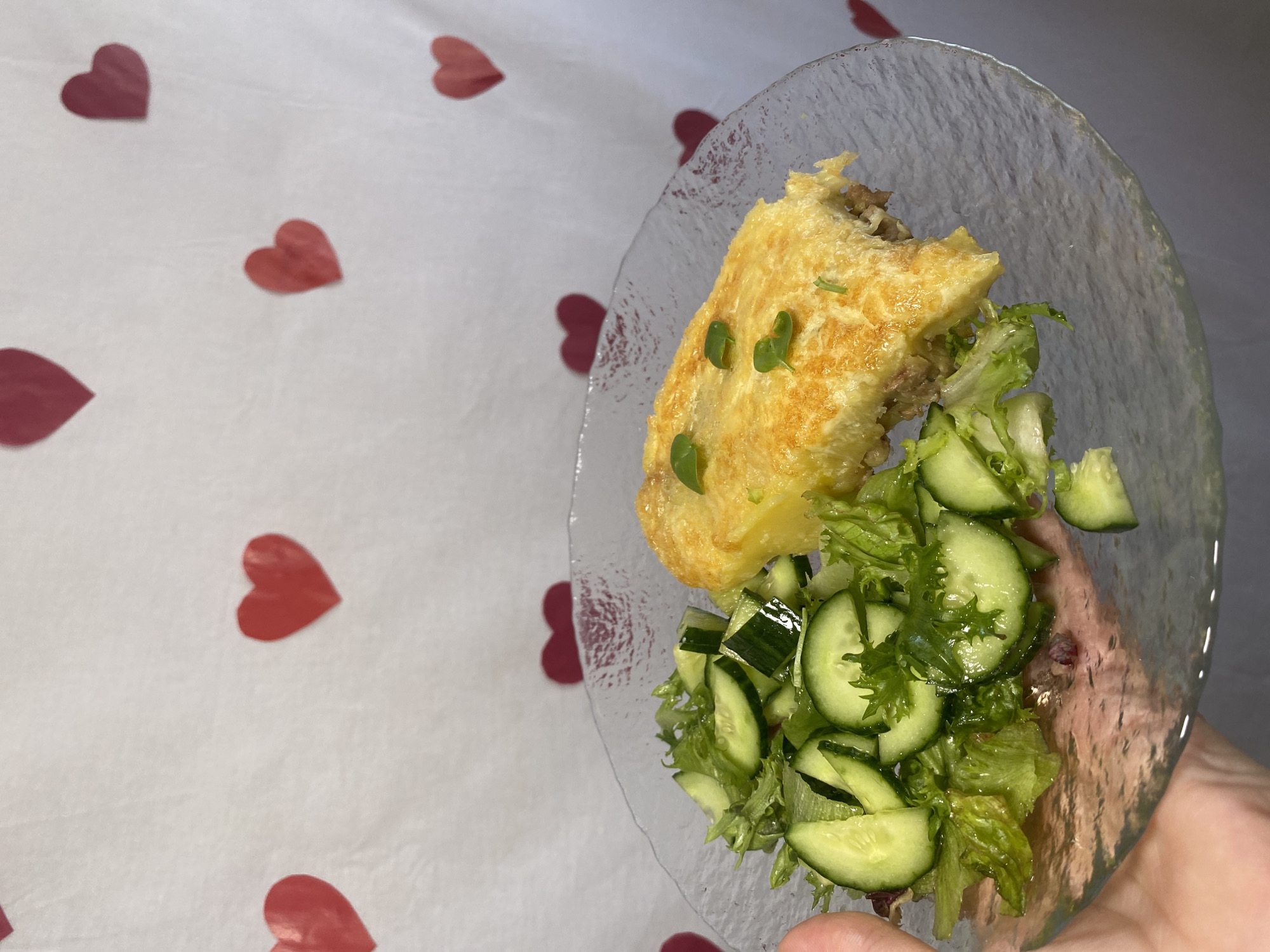 A plate with a portion of omelet and cucumber salad is held in a person's hand. The background is a white surface with scattered red heart shapes. The location is Chaikovsky, Russia.