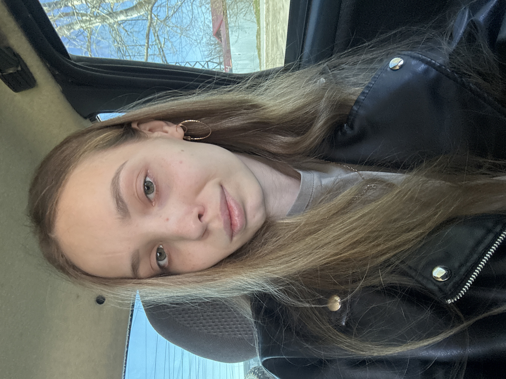 A fair-skinned female with light brown hair is in the driver's seat of a car. She is wearing a gray t-shirt and a black leather jacket. Her hair is long and partially covers the jacket. She is wearing gold hoop earrings. The car's interior is visible, with a gray headrest and black trim. Outside the car window, trees and buildings are visible under a clear, blue sky. The location is Chaikovsky, Russia.