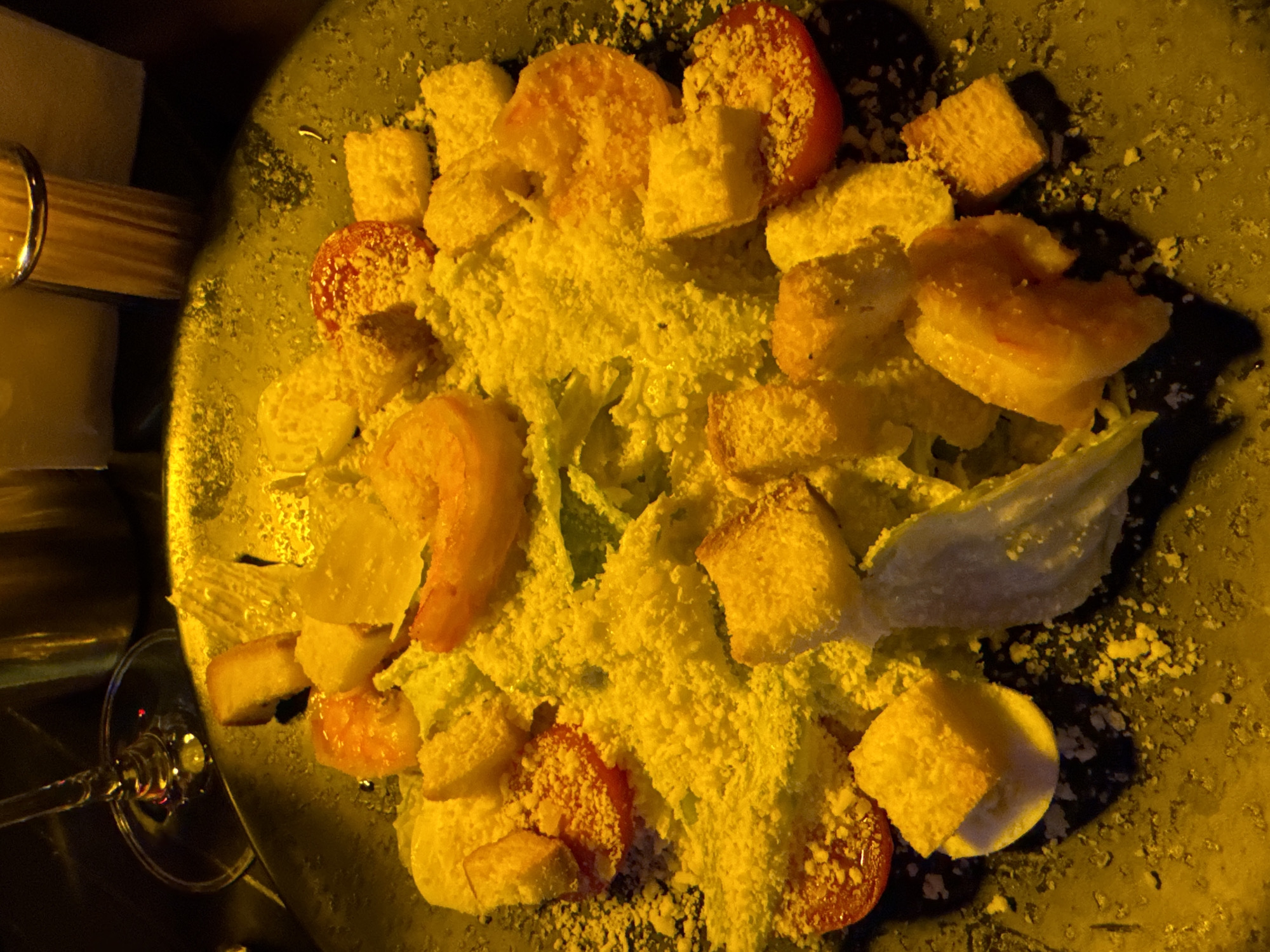 The image presents a top-down view of a Caesar salad served on a dark-colored plate, possibly ceramic or stoneware. The salad consists of romaine lettuce, croutons, sliced tomatoes, and appears to be heavily dressed with grated cheese. The lighting is warm, casting an overall yellow hue on the scene. To the left of the plate, a partially visible wine glass and a cylindrical object, potentially a napkin holder or part of the table setting, can be seen. The background is dark, suggesting the setting is indoors, perhaps a restaurant or dining establishment. The specific city is not discernible from the image.