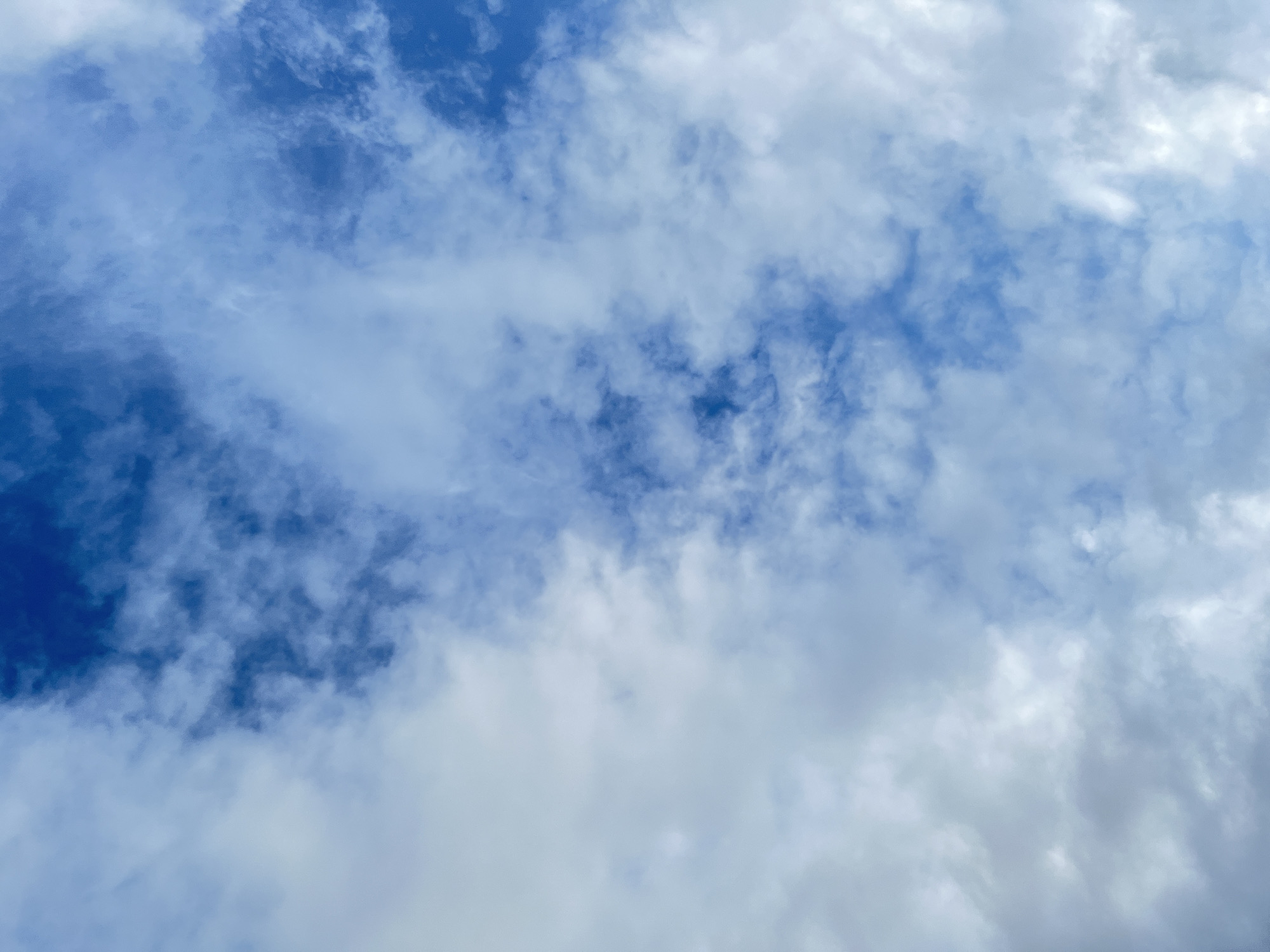 The image displays an expansive view of the sky over Lhokseumawe, Indonesia. The scene is dominated by a mixture of blue sky and scattered white to light gray clouds. Patches of clear, deep blue sky are visible, particularly in the upper right and along a central vertical band, contrasting with the more diffuse, lighter blue areas where thinner clouds are present. The clouds themselves are amorphous and varied in density, ranging from translucent, wispy formations to more opaque, brighter white accumulations. No people, objects, or ground features are discernible within the frame, which focuses exclusively on the atmospheric conditions.