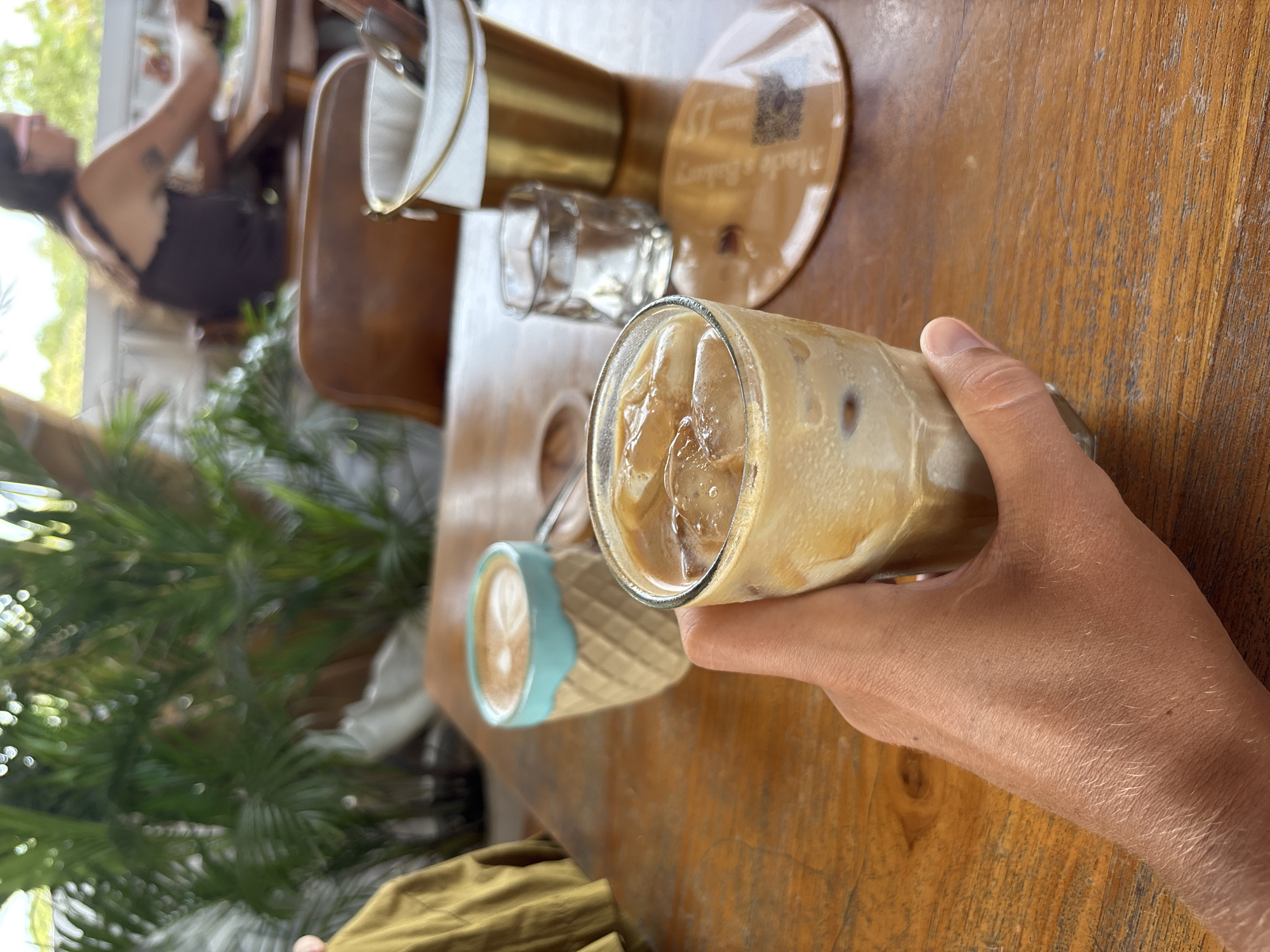 The image captures a casual cafe scene, likely outdoors or in a bright, open-air setting during the daytime. The perspective is from an individual's point of view, holding a beverage.
In the foreground, a fair-skinned hand firmly grips a clear glass filled with iced coffee. The coffee is a creamy, light brown color, containing numerous ice cubes. To the left of the iced coffee, a light blue and beige patterned cup, possibly ceramic, holds another beverage with visible latte art on its surface.
The objects on the wooden table include a clear empty glass, and part of a golden pour-over coffee apparatus. A round, light brown coaster is visible, with faint text that reads "Made a Bakery" and a small QR code. The table itself is made of light-colored wood, showing some wear and grain.
In the blurred background, lush green foliage, resembling tropical plants, frames the scene, suggesting a natural and relaxed environment. Also in the background, a person with dark hair, wearing a dark top, is partially visible and heavily out of focus, seated at another table or counter.
The image depicts a moment of leisure, with someone enjoying a cold drink at a cafe. The setting, with iced beverages and tropical greenery, aligns with the provided location context of Kuta Selatan, Indonesia.