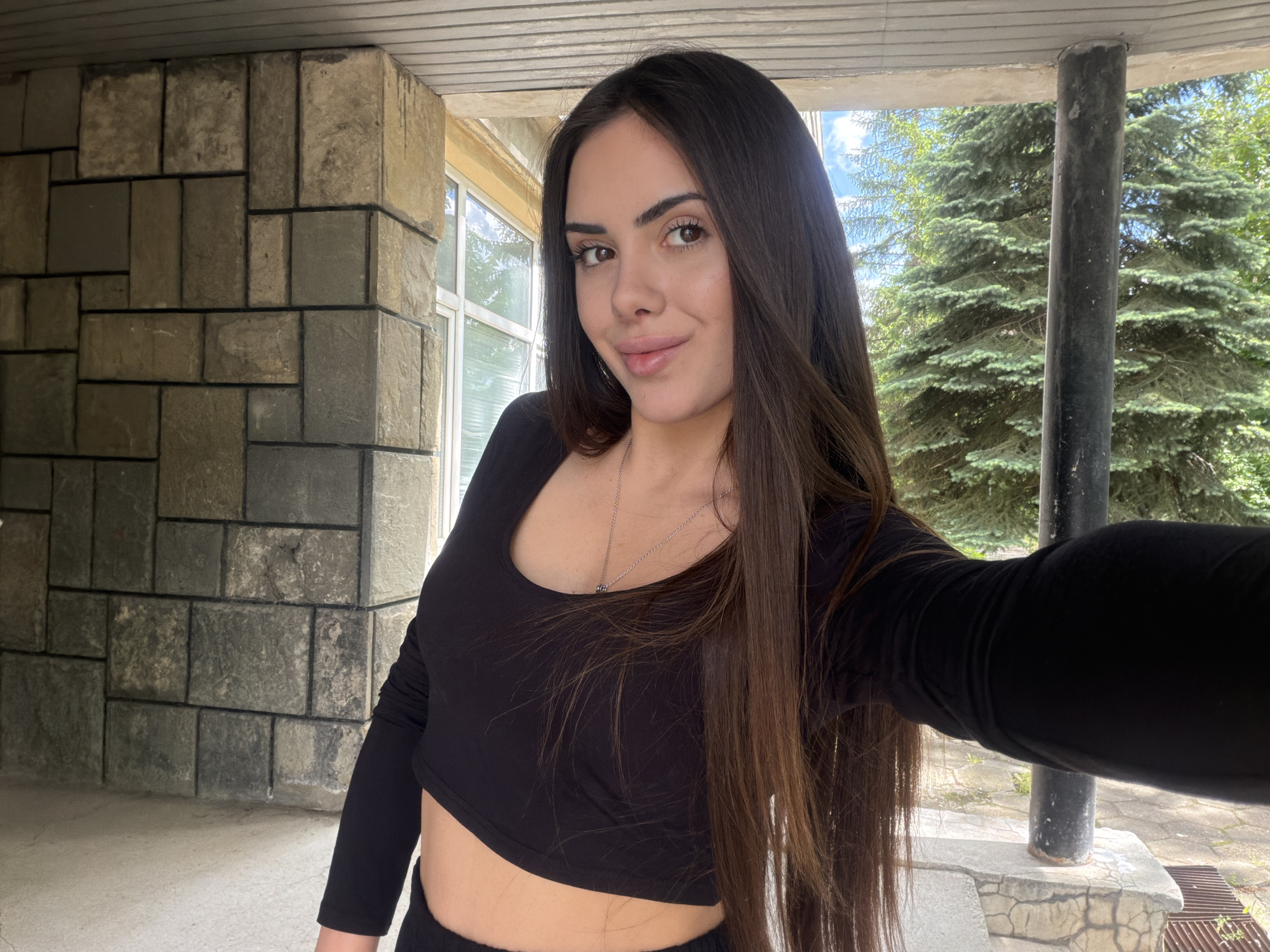 A woman is taking a selfie outdoors. She has long, dark hair and is wearing a black crop top. Behind her is a building constructed of large, rough-cut stone blocks and a window. Further back, trees and sky are visible. The specific location cannot be determined without additional information. This location is not known to be within the city of Sofia, Bulgaria.