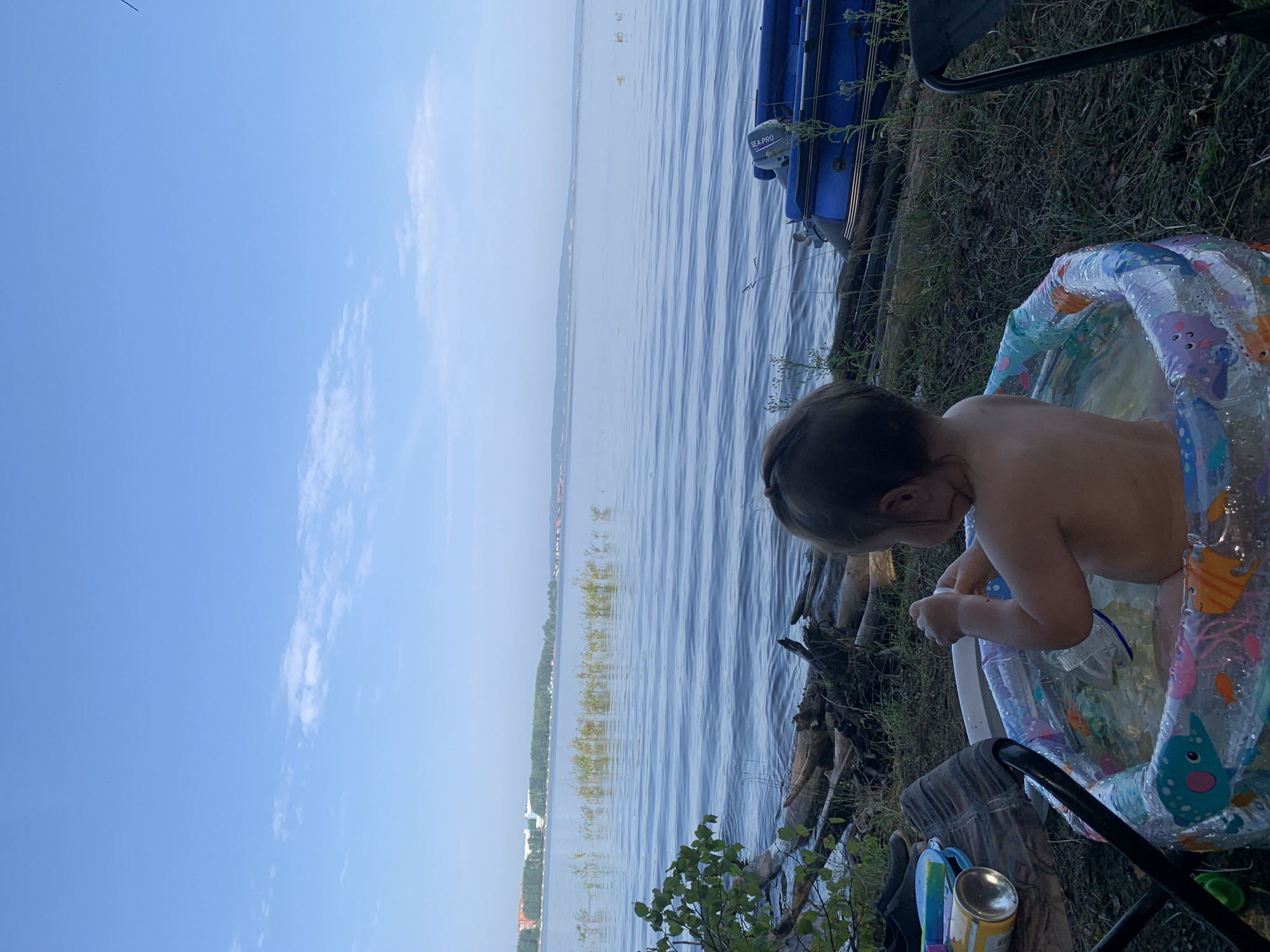 In this image, a young child, seen from behind, is sitting in a colorful inflatable kiddie pool filled with water. The child is partially submerged and appears to be playing with something in the water. The pool is set up on grassy ground near a body of water that stretches to the horizon. A blue inflatable boat with "SEA-PRO" printed on its engine is visible to the right of the kiddie pool. The scene suggests an outdoor recreational setting, possibly a lakeside or riverbank. The sky is a clear, light blue with some wispy clouds, indicating fair weather and likely daytime, possibly late afternoon or early evening due to the lighting. The overall mood is peaceful and relaxed, highlighting a moment of childhood enjoyment in a natural environment.