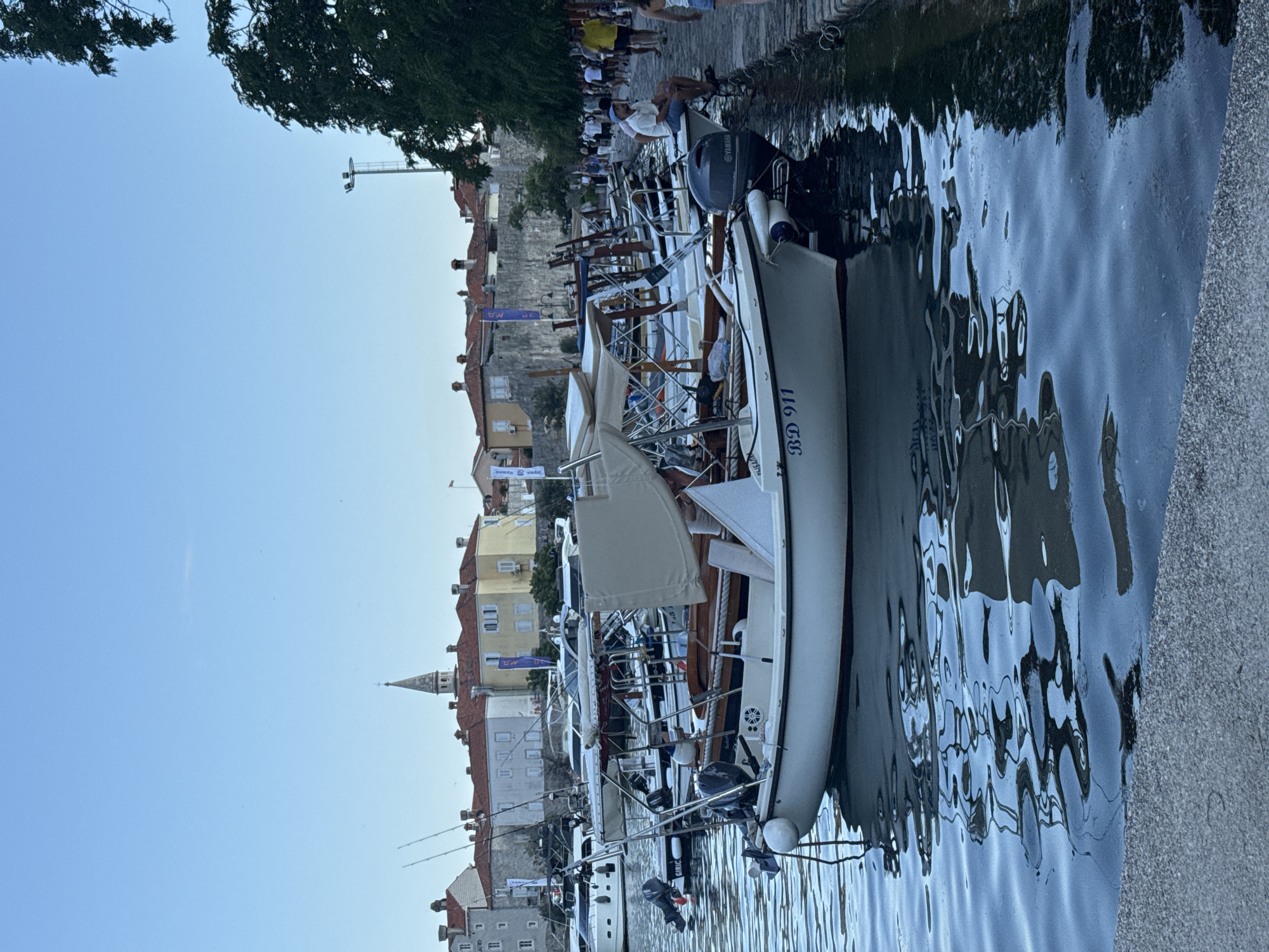 The image shows a harbor in Budva, Montenegro, with several small boats docked along a stone quay. In the foreground, a white motorboat with the registration "BD 911" is prominent. The boat has a canvas canopy and a Yamaha outboard motor. Other boats are moored behind it, and further out, larger yachts are visible.
In the background, the historic Old Town of Budva can be seen, characterized by its red-tiled roofs, stone walls, and a church spire. A tree overhangs the right side of the image, partially obscuring the view of the town.
Along the quay, several people are walking and sitting. One person is seated on the edge of the quay, near a boat, facing the water. The water is calm with reflections of the boats and the sky. The overall scene depicts a picturesque coastal town with a bustling harbor.