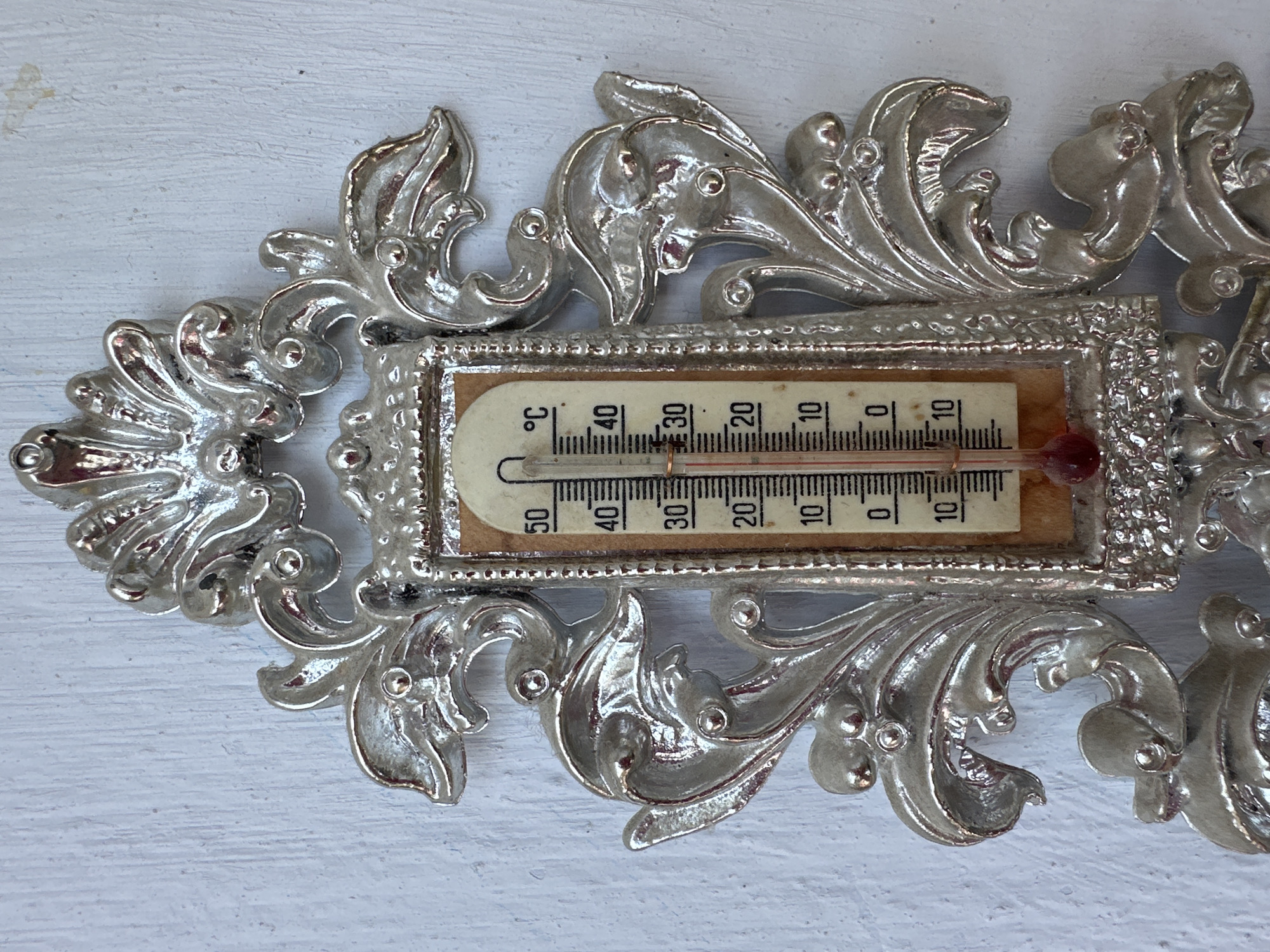 A close-up, high-angle, eye-level shot captures an ornate, silver-colored thermometer mounted on a textured, white wall. The thermometer, encased in a decorative frame with floral and scrolling motifs, displays a temperature reading of approximately 25 degrees Celsius. The thermometer's glass tube contains a red liquid that has risen to just below the 30-degree mark, with clear markings for every 10 degrees and finer divisions in between. The wall's surface exhibits a subtle, uneven texture, suggesting it might be painted wood or plaster. The overall scene is static and devoid of any human presence or activity, presenting a solitary artifact against a neutral backdrop. The location is identified as Peremyshliany, Ukraine.