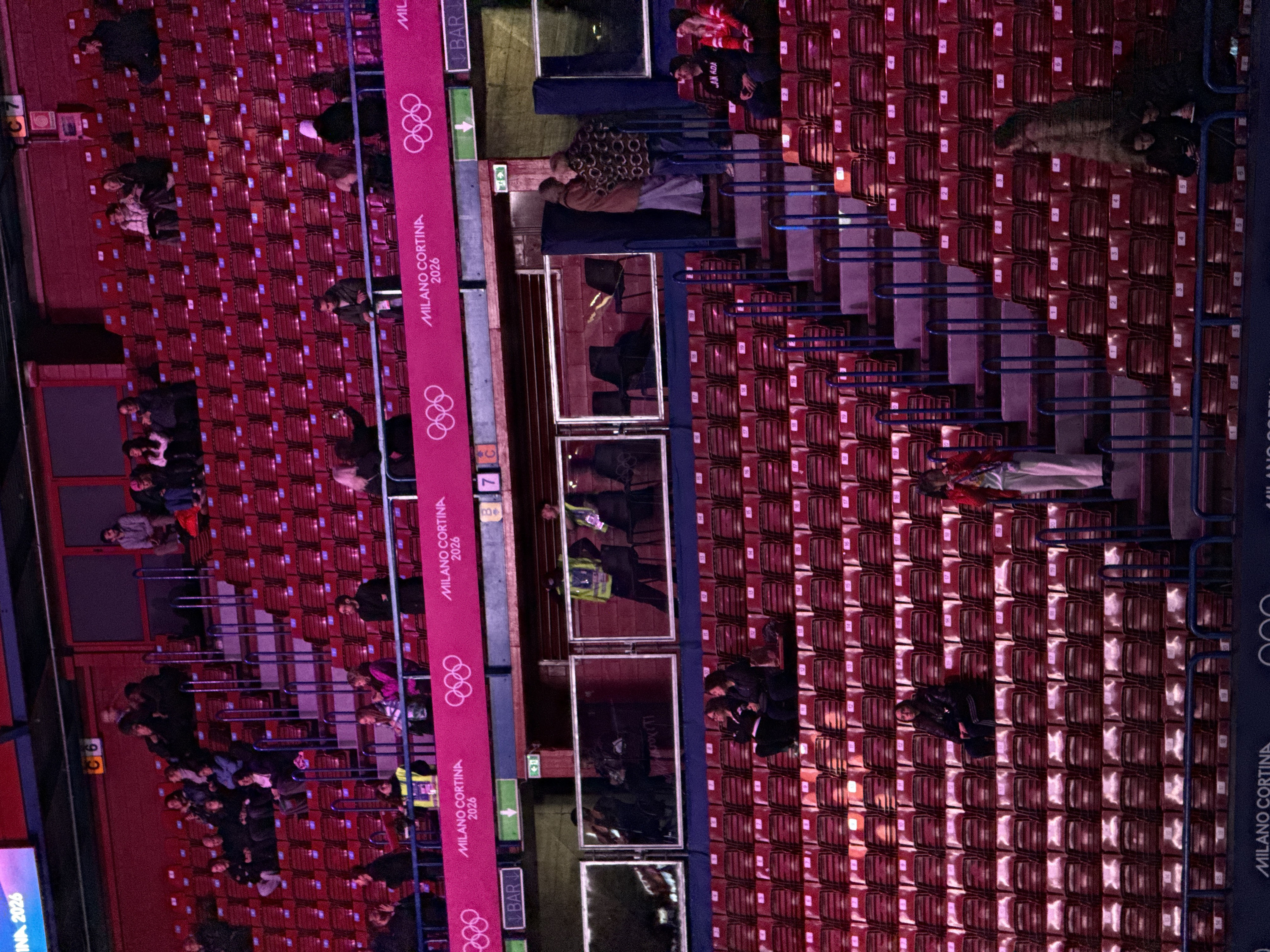 The image captures an indoor stadium or arena, predominantly filled with rows of empty red seats, suggesting it's either before an event, during a break, or a sparsely attended section. The perspective is from above, looking down on the seating areas and a walkway or platform below.
Numerous people are scattered throughout the scene. Some are seated in small groups, chatting or looking towards an unseen event. Others are standing in the walkways or near the railings. In the upper right section, a person is visible wearing a red jacket with "CANADA" written on the back. Further down in the right seating area, another individual appears to be resting or sleeping across several seats. In the lower left, a group of people is visible near a railing. In the center-right, staff members, possibly security or ushers, are wearing high-visibility vests and are seen behind glass partitions.
The seats are numbered, with visible numbers like 1, 2, 3, 4, 5, and 6 on the backrests, indicating seat and row assignments. Blue metal railings divide the seating sections and provide support.
Prominently displayed throughout the image are vertical pink banners featuring the Olympic rings logo and the text "MILANO CORTINA 2026", strongly indicating a connection to the upcoming Winter Olympic Games in Milan and Cortina, Italy. Other signs include a "BAR" sign with an arrow pointing left, and section indicators such as "C7" and "C6". A green sign with a white arrow indicates an exit.
The overall lighting is artificial and somewhat dim, consistent with an indoor stadium environment during an evening event or practice. The scene is relatively quiet, without the visible energy of a fully packed stadium.