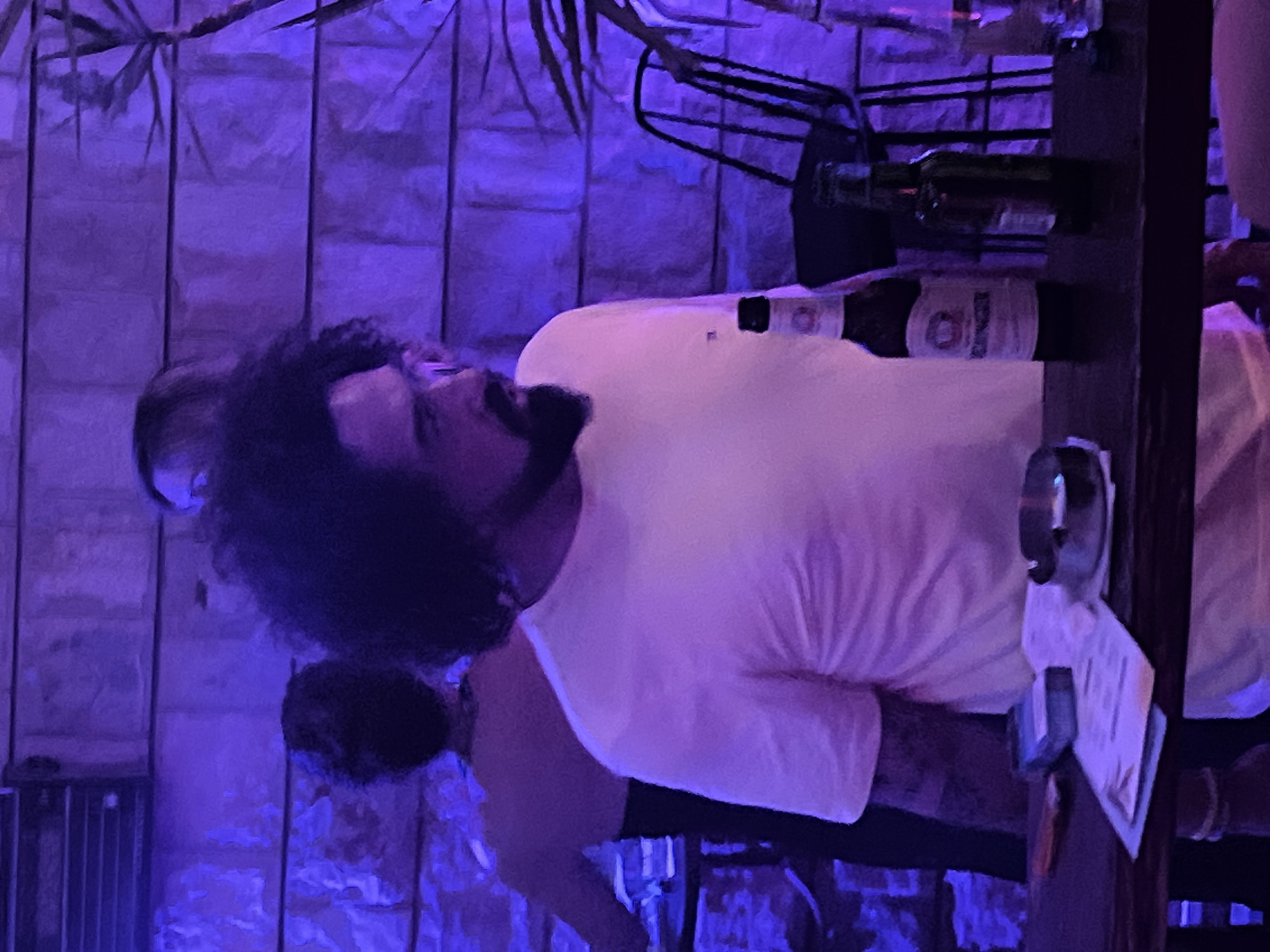 The image depicts a dimly lit indoor scene, possibly a bar or restaurant, bathed in purple ambient light. In the foreground, a man with curly dark hair and a beard, wearing a light-colored t-shirt, stands at a dark wooden counter. He is looking to his left. On the counter, there are two beer bottles, one with a white label and the other dark green, an ashtray with a cigarette butt, and what appears to be a pack of cigarettes and some papers. In the background, a woman with dark hair is visible from the back, wearing a black dress. Another person's head is partially visible behind the man in the foreground. The wall behind them is made of textured stone blocks. The scene suggests a relaxed, social atmosphere.