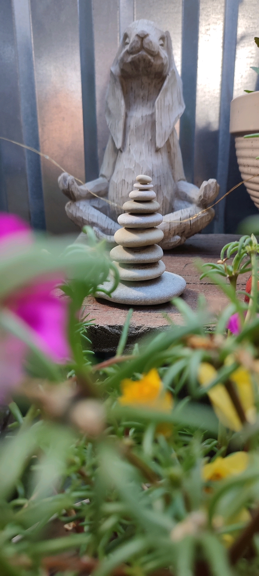 In Belaya Kalitva, Russia, an outdoor scene features a stone statue of a rabbit in a meditating pose, positioned behind a stack of smooth, grey stones arranged in a cairn. The backdrop is a corrugated metal wall. In the foreground, out-of-focus pink and yellow flowers and green foliage create a soft bokeh effect. The lighting suggests it is daytime.