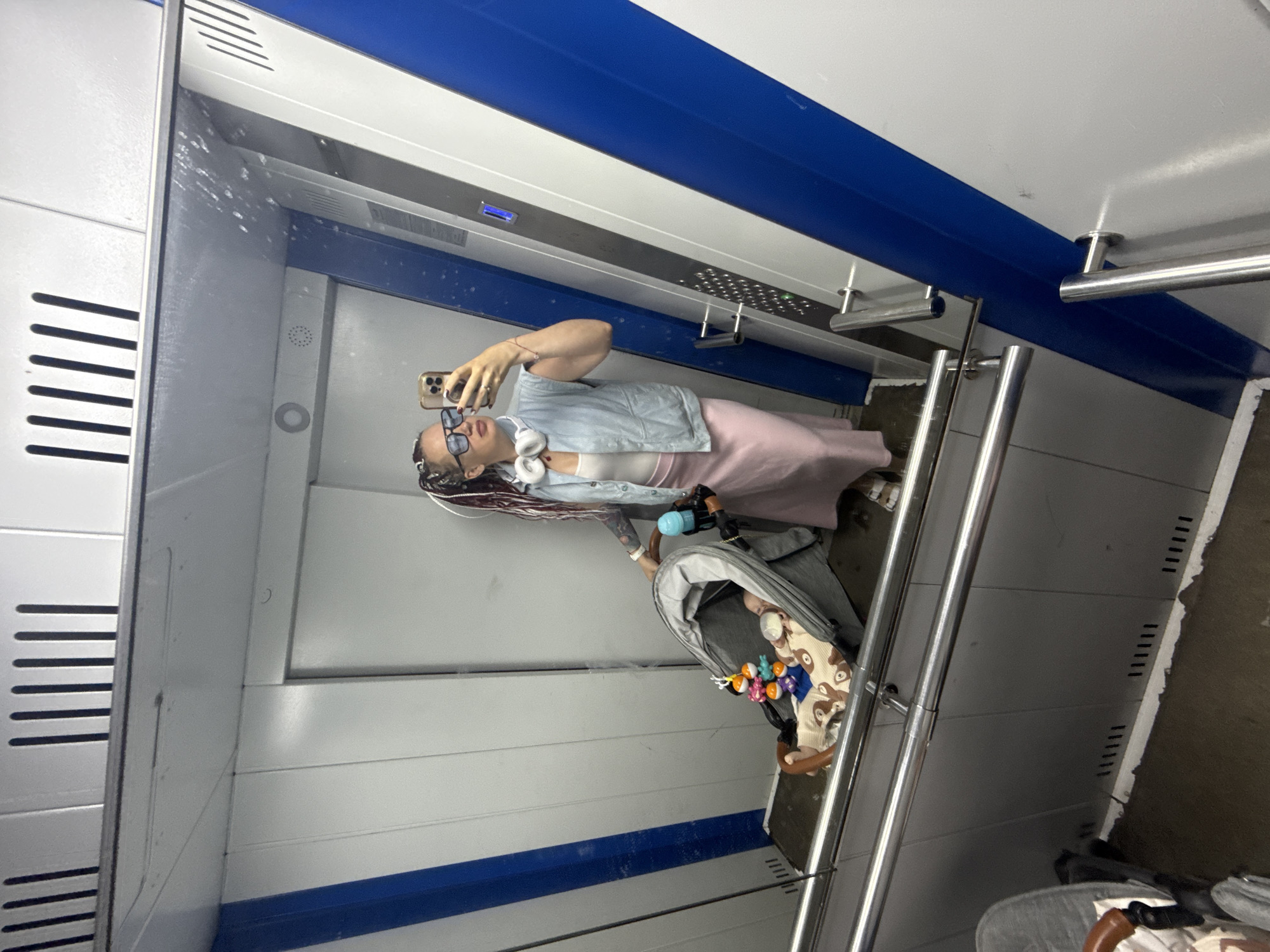 An image, rotated 90 degrees clockwise, captures a mirror selfie taken inside what appears to be a modern public transport vehicle or elevator, possibly in Moscow, Russia.
The primary subject is a young adult woman, reflected in a large mirror. She is holding an iPhone in her right hand, angled to take the picture. She wears dark-rimmed eyeglasses, a light blue denim vest over a white top, and a pink skirt. White headphones are around her neck, and her hair is styled in long braids or dreadlocks. A tattoo is visible on her left forearm.
Beside her, to her left in the reflection, is a baby stroller with an infant inside. The baby is wearing a patterned garment and appears to be holding a baby bottle. Colorful toys are attached to the stroller's bar.
The interior of the space features clean, white panels with a prominent horizontal blue stripe running across. There are multiple metallic handrails and support bars. On the left side of the reflection, a panel with horizontal slats is visible, suggesting a ventilation system or design element typical of a train car. The mirror itself has some visible smudges. The floor area, partly obscured, shows grey panels with small, rectangular vents. No discernible text is visible within the frame.