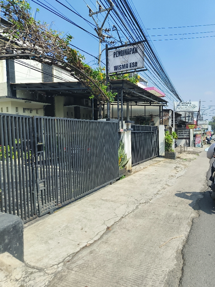 The image shows the exterior of a lodging establishment called "Wisma Esa" in Ngamprah, Indonesia, located at coordinates -6.865 latitude and 107.512 longitude. The establishment has a black metal gate and a sign advertising "Penginapan" (Lodging) and "Wisma Esa." The gate is closed. To the right of the gate, there is a paved sidewalk. A motorcycle is parked on the street, partially out of frame. Several power lines are seen overhead. The scene appears to be taken in the daytime, with clear skies and a bright light. The image was taken from a perspective facing the building, with the camera positioned slightly to the left of center.
