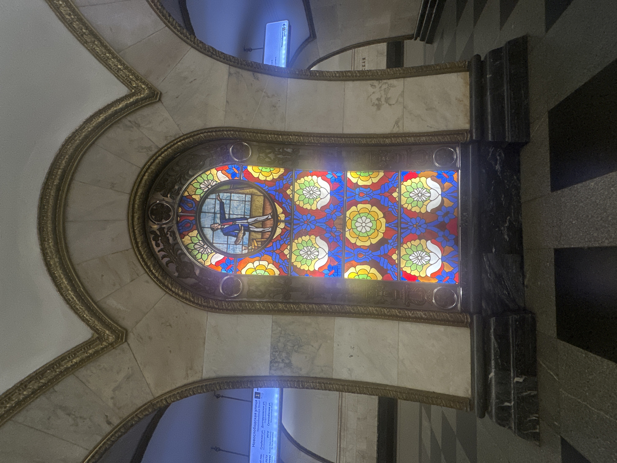 The image depicts a stained-glass window in a Moscow Metro station. The window is set within a decorative archway with marble and ornate gold trim. The stained glass features a central circular depiction of a man in a blue jacket and red scarf holding a palette and brush, with a framed window behind him. Below this scene, geometric floral patterns in vibrant colors like red, blue, yellow, green, and white are repeated. The window is illuminated from behind, making the colors stand out. The station's architecture includes marble walls, arched passageways visible on the left and right, and a checkered floor made of dark and light gray tiles. Signs in Russian are visible in the background, indicating the station name is "Novoslobodskaya Street." The overall impression is one of grandeur and artistry within a public transit space.