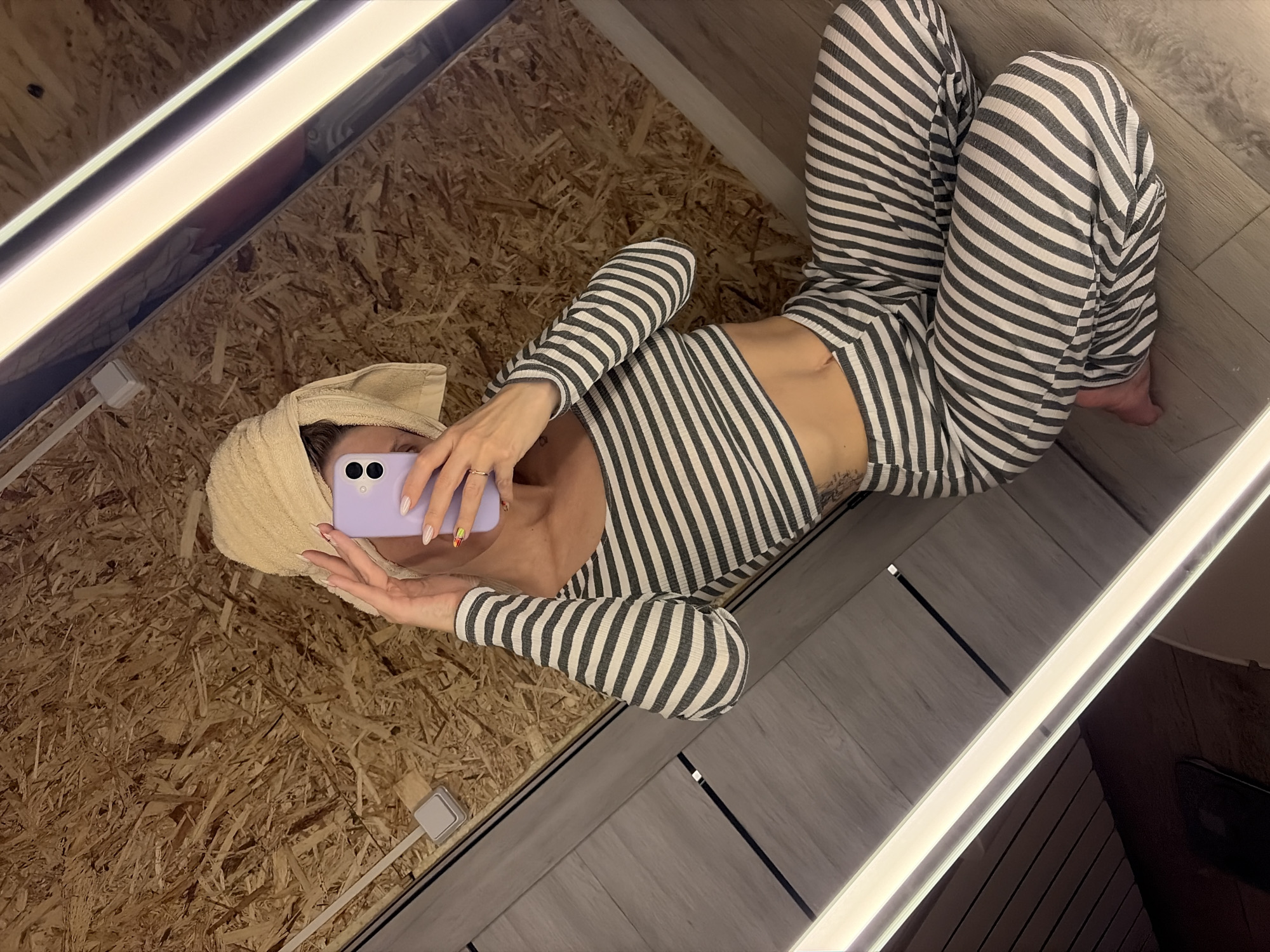 This image depicts an interior scene in Chaikovsky, Russia, captured as a mirror selfie. An individual, whose face is obscured by a light purple smartphone held with both hands, is seated cross-legged on a light grey wood-plank patterned floor. A light beige towel is wrapped around the person's head. They are wearing a two-piece ribbed loungewear set, featuring a long-sleeved crop top and wide-leg pants, both displaying alternating grey and white horizontal stripes. Visible on the person's left hand is a gold-colored ring on the ring finger, and both hands exhibit manicured fingernails with varied polish designs. A dark tattoo is partially visible on the left side of the person's torso/hip area. The background reflected in the mirror shows a wall constructed from light brown, textured wood composite material, and a light grey square electrical fixture is mounted on this wall. Two long, vertical, illuminated LED strip lights flank the mirror, providing primary light.