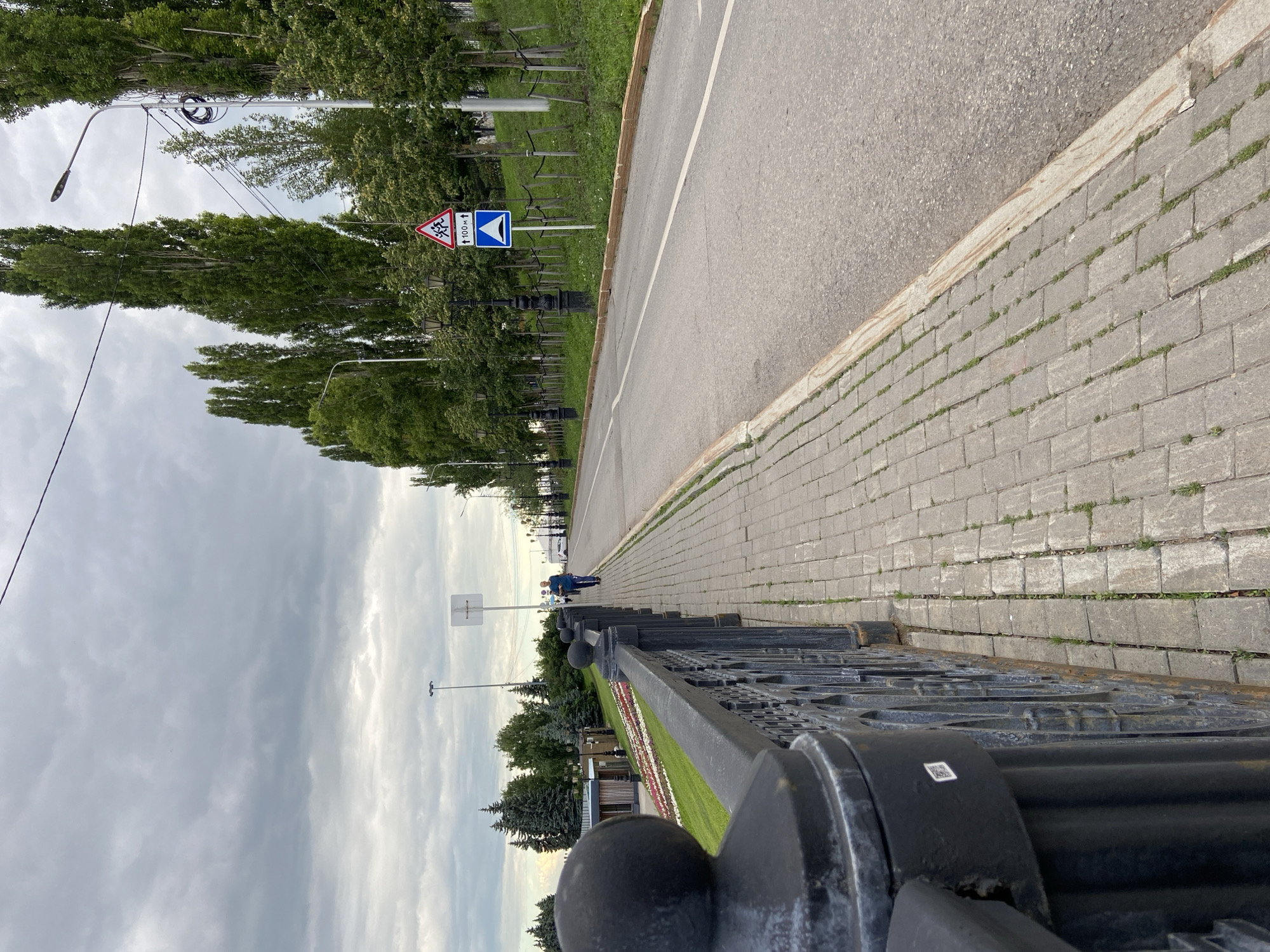The image depicts a view of a street lined with trees and a paved walkway. In the foreground, a decorative black metal fence runs along the edge of the walkway. The walkway is paved with grey bricks, and beyond it is a paved road with a white line marking. Further in the distance, there are more trees and some buildings, including what appears to be a small pavilion. A few street lights and utility poles are also visible.
There are two people walking on the walkway in the distance. One person is wearing a blue jacket and appears to be walking towards the viewer. The other person is further away and is partially obscured by trees.
Two road signs are visible on the left side of the road. One is a red and white triangular sign with a symbol depicting multiple cars or obstacles, likely indicating a hazard or uneven road surface. The other is a blue rectangular sign with a white symbol of a speed bump or hump. The triangular sign also has a white arrow pointing upwards and the text "100м" which means "100 meters" in Russian, suggesting a hazard or caution within that distance.
The sky is overcast with clouds, suggesting a cloudy or possibly rainy day. The lighting is diffused. The time of day appears to be late afternoon or early evening, given the angle of the light and the overall mood. The presence of the Russian text on the sign and the overall urban park-like setting are consistent with the provided location context of Ufa, Russia. The scene conveys a sense of calm and ordinary urban life.