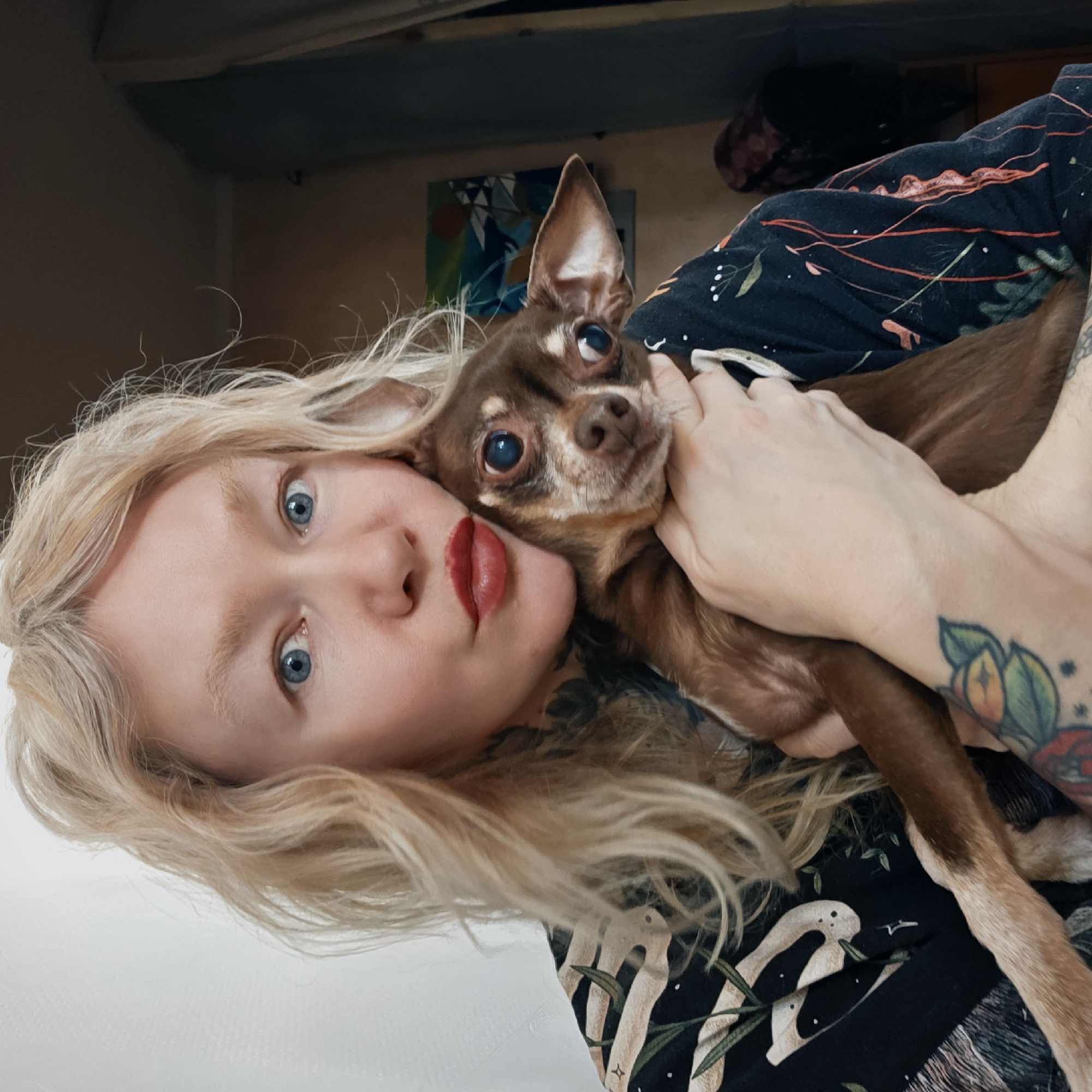 A woman with blonde hair and red lipstick is holding a brown chihuahua-type dog in her arms. The woman has blue eyes and tattoos on her arm. She is wearing a dark-colored shirt with a light-colored pattern. The dog has large, expressive eyes. In the background, there is a painting on the wall and what appears to be a storage space. The location is not provided.