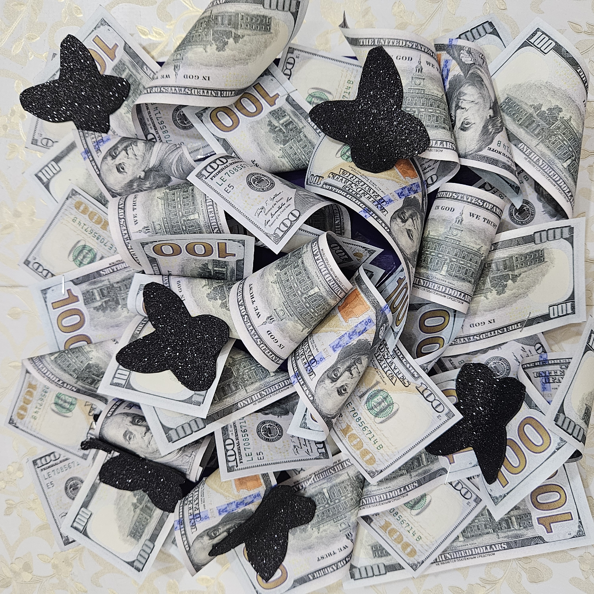 The image displays a close-up, overhead view of a pile of US hundred-dollar bills. The bills are crumpled and partially rolled, creating a textured surface. Several black, glittery butterfly-shaped decorations are scattered amongst the money. The background appears to be a light-colored fabric with a subtle floral pattern. There are no people or any elements indicating a specific city or location within Andijan City, Uzbekistan. The scene is static and focused solely on the money and the decorative butterflies.