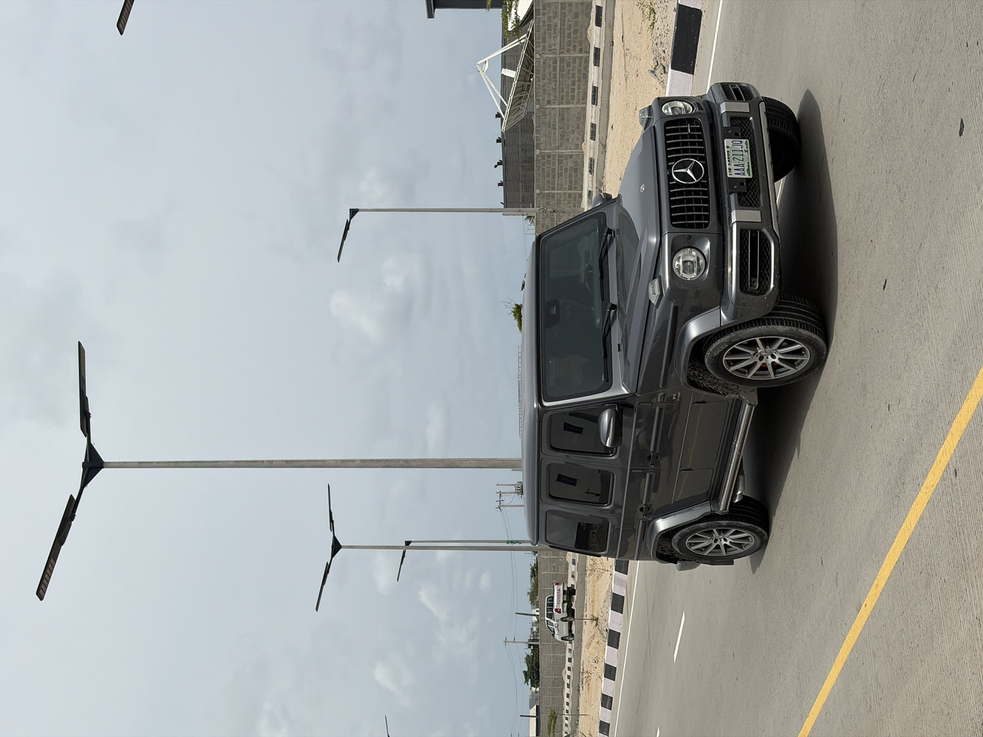 The image shows a gray Mercedes-Benz G-Class SUV parked on a paved road, likely in Alaguntan, Nigeria. The car is angled slightly towards the right side of the frame. The license plate reads "AAA-211JO" and indicates it is from Lagos, Nigeria. The car is parked near a curb marked with black and white stripes. The road also has a yellow lane marking. In the background, there are modern streetlights with a unique three-pronged design. There is a building under construction with exposed brick walls and a partially erected roof structure. A white Toyota pickup truck is visible in the background. The sky is overcast, suggesting a cloudy day.