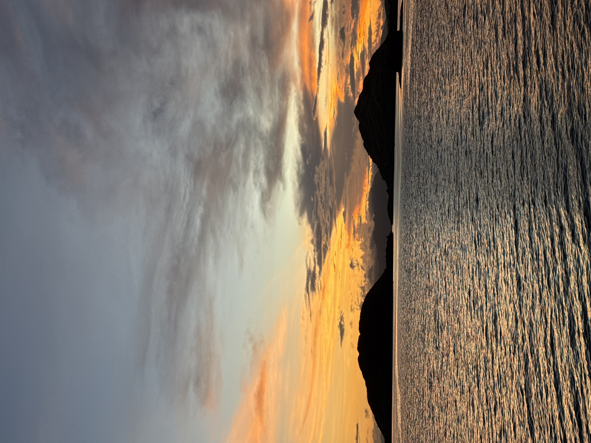 The image depicts a coastal scene at sunset. On the right, dark, rippled water fills a large portion of the frame, reflecting the light of the setting sun. In the middle, a dark silhouette of a mountain or landmass separates the water from the sky. The sky features dramatic clouds with hues of orange and yellow near the horizon, transitioning to grey and white higher up. The location is possibly in Indonesia, based on the context provided in the prompt.