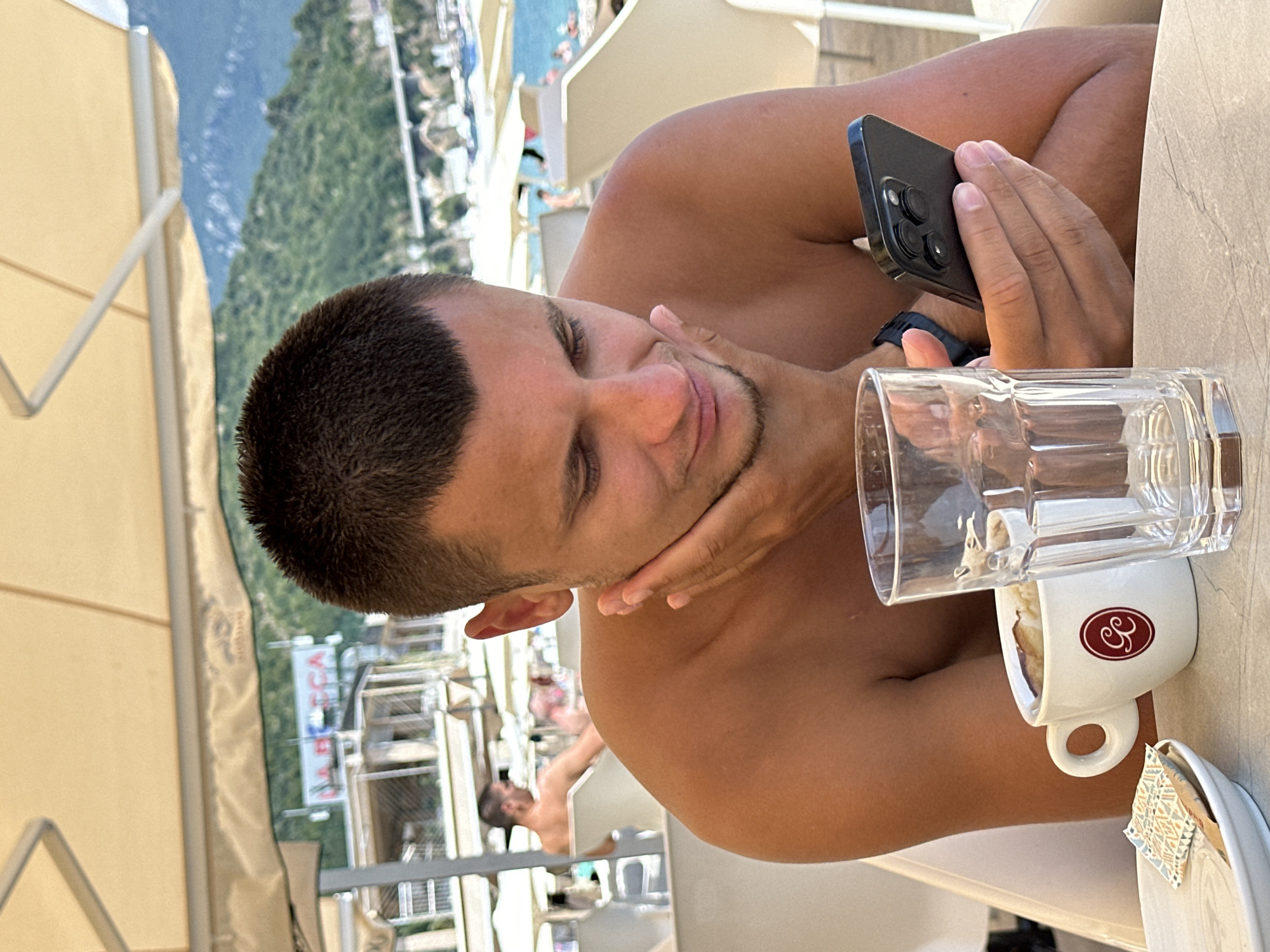 A young man with short brown hair and a light beard is sitting at a table outdoors, holding a black smartphone in his right hand and resting his chin on his left hand. He is looking down at his phone. In front of him, there is a white cup with a red logo, a clear glass, a plate, and a packet of sugar. The man is shirtless. In the background, there are beach umbrellas, lounge chairs, people on a beach, and a body of water with boats and hills beyond. The scene appears to be at a cafe or restaurant near a beach in Budva, Montenegro, likely on the Adriatic coast given the mountainous landscape and the type of coastal development.