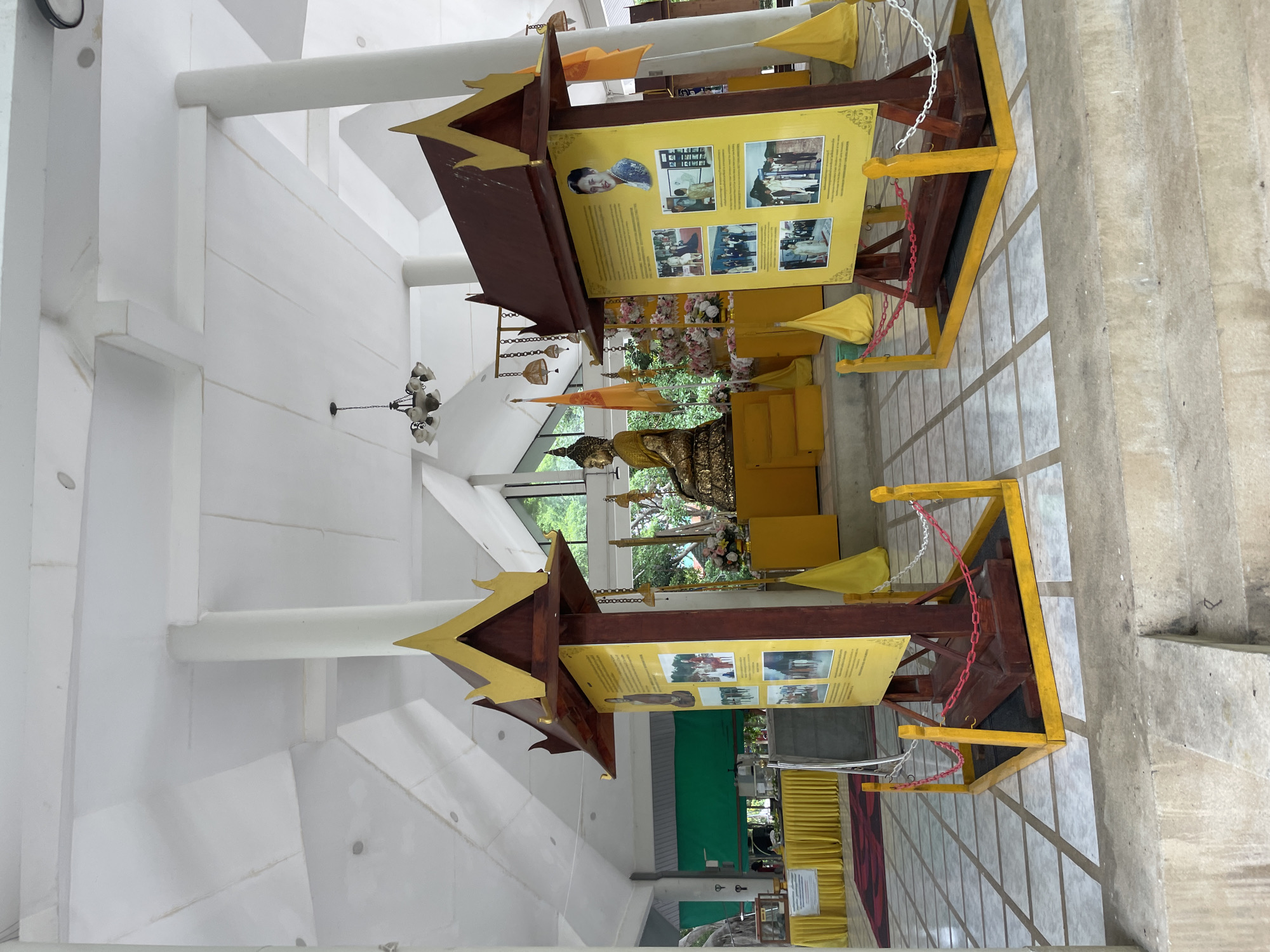 The image depicts the interior of a Buddhist temple or shrine, likely located in Na Chom Thian, Thailand. Two large display boards, topped with ornate wooden structures, stand prominently in the foreground, separated by a patterned tiled floor. These boards feature framed photographs and text, with the board on the right showcasing a portrait of a woman and various historical images.
Behind the display boards, a seated golden Buddha statue is visible, adorned with gold leaf. It is placed on a raised platform surrounded by yellow fabric decorations and floral arrangements. Several yellow fabric flags and hanging decorative bells are also present.
The ceiling is high and white, supported by white pillars. Natural light streams in from windows at the rear of the structure, revealing greenery outside. In the background, to the left, a yellow draped table with items on display can be seen, and further to the left, a green fabric curtain hangs. The overall scene suggests a place of worship and historical exhibition.