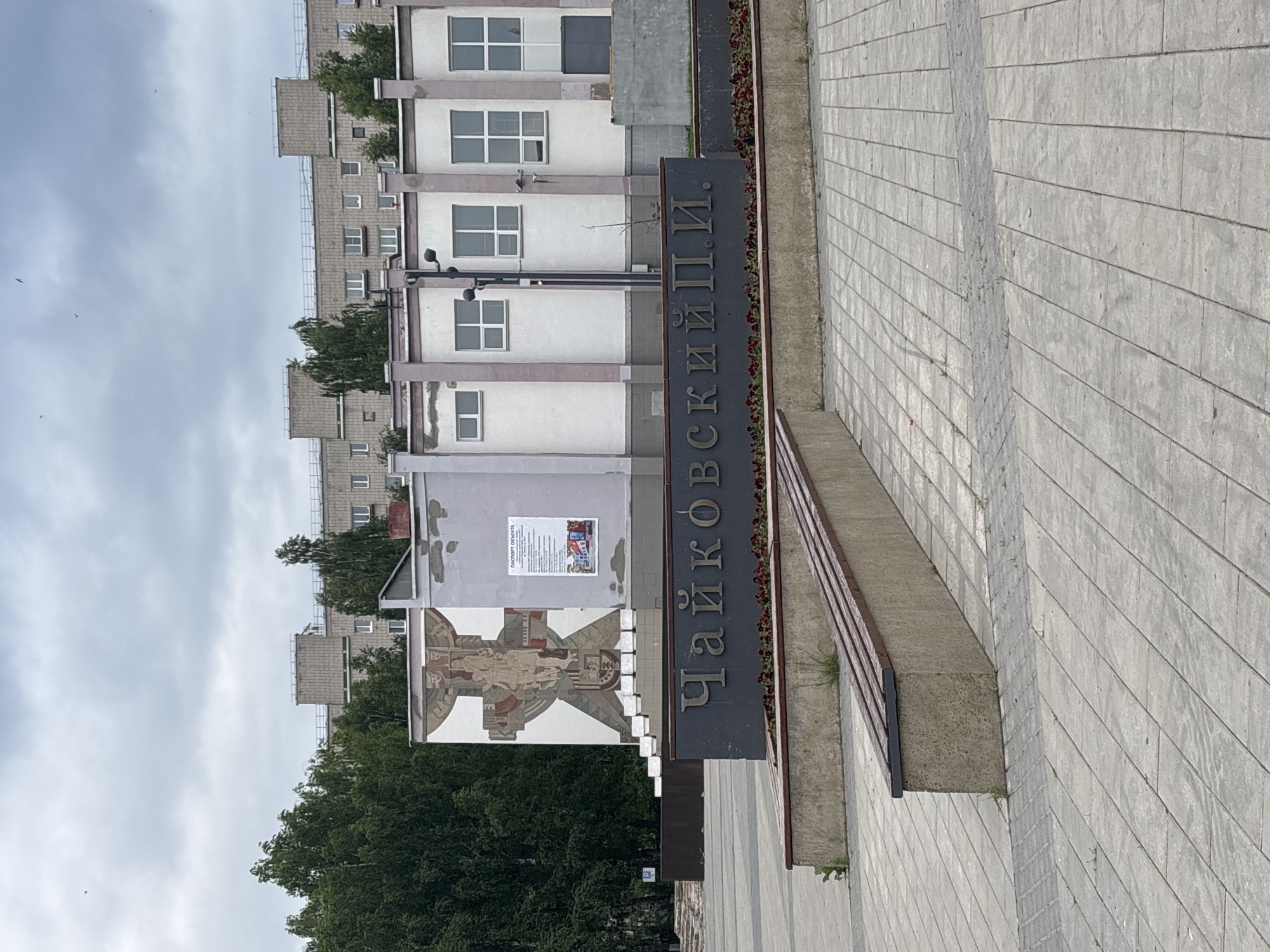 A wide shot shows a public square with a monument in the foreground. The monument is a dark gray slab with embossed gold lettering that reads "Чайковский П.И." ("Tchaikovsky P.I."). Behind the monument is a low, concrete bench with wooden slats. The square is paved with gray interlocking tiles. In the background, a building with a mural on its left side is visible. The building has light purple accents and several windows. Trees and a cloudy sky complete the scene.