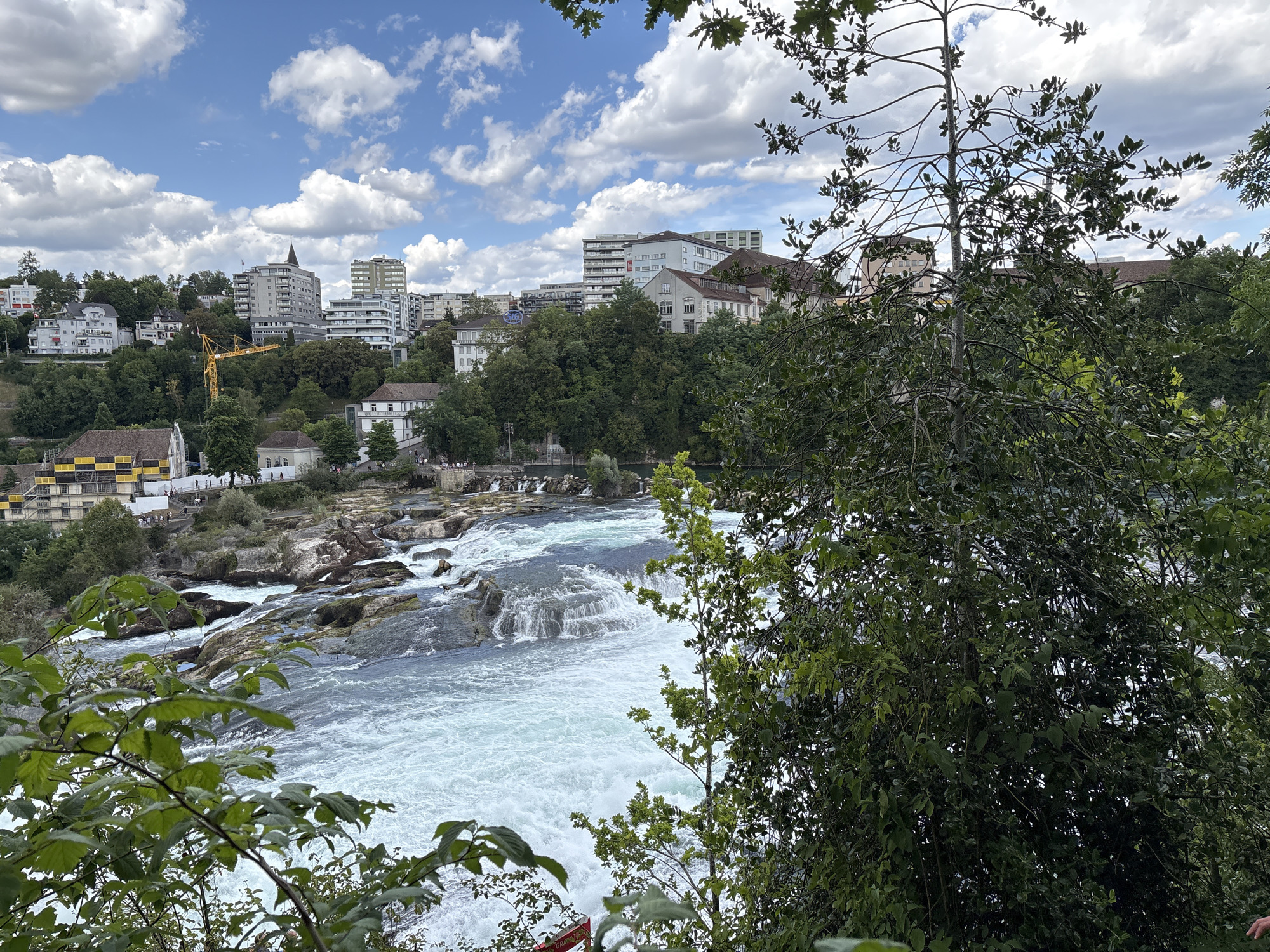 The image captures a wide view of the Rhine Falls, a stunning waterfall located near Dachsen, Switzerland. The falls are a dominant feature, with powerful white water cascading over jagged rocks. The surrounding landscape is lush and green, with dense trees framing the scene. In the background, a cityscape is visible, with modern buildings and older structures nestled on a hillside. A yellow construction crane is also present, suggesting ongoing development in the area. The sky is a vibrant blue, dotted with fluffy white clouds, indicating fair weather. The overall impression is one of natural beauty and urban life coexisting.