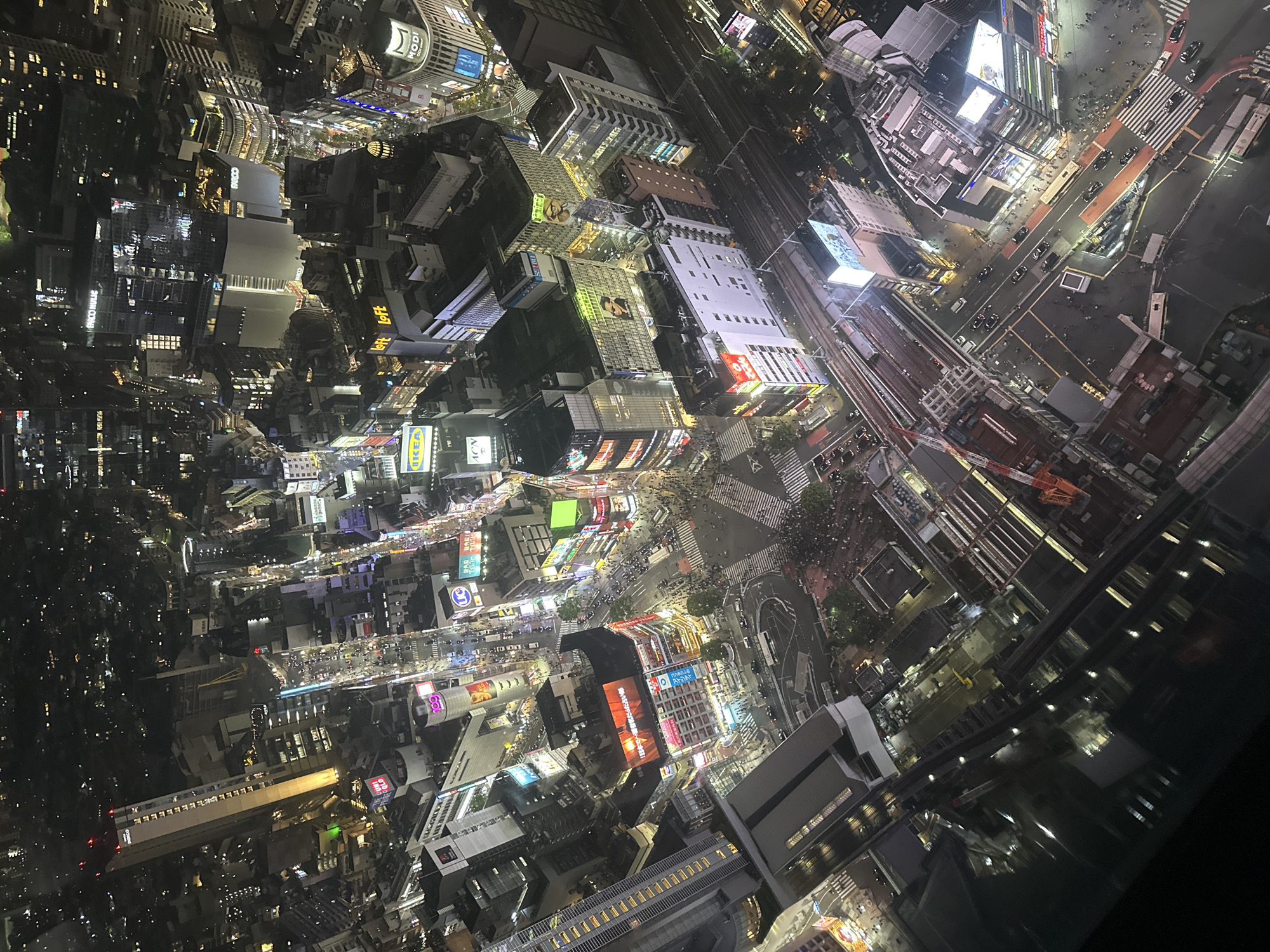 The image captures an aerial nighttime view of Shibuya, Japan, characterized by densely packed buildings illuminated with an array of lights. A significant pedestrian scramble crossing, crowded with people, is prominently featured. Major buildings and structures include train tracks and stations, contributing to the area's transportation infrastructure. The general environment suggests a bustling urban atmosphere at night.