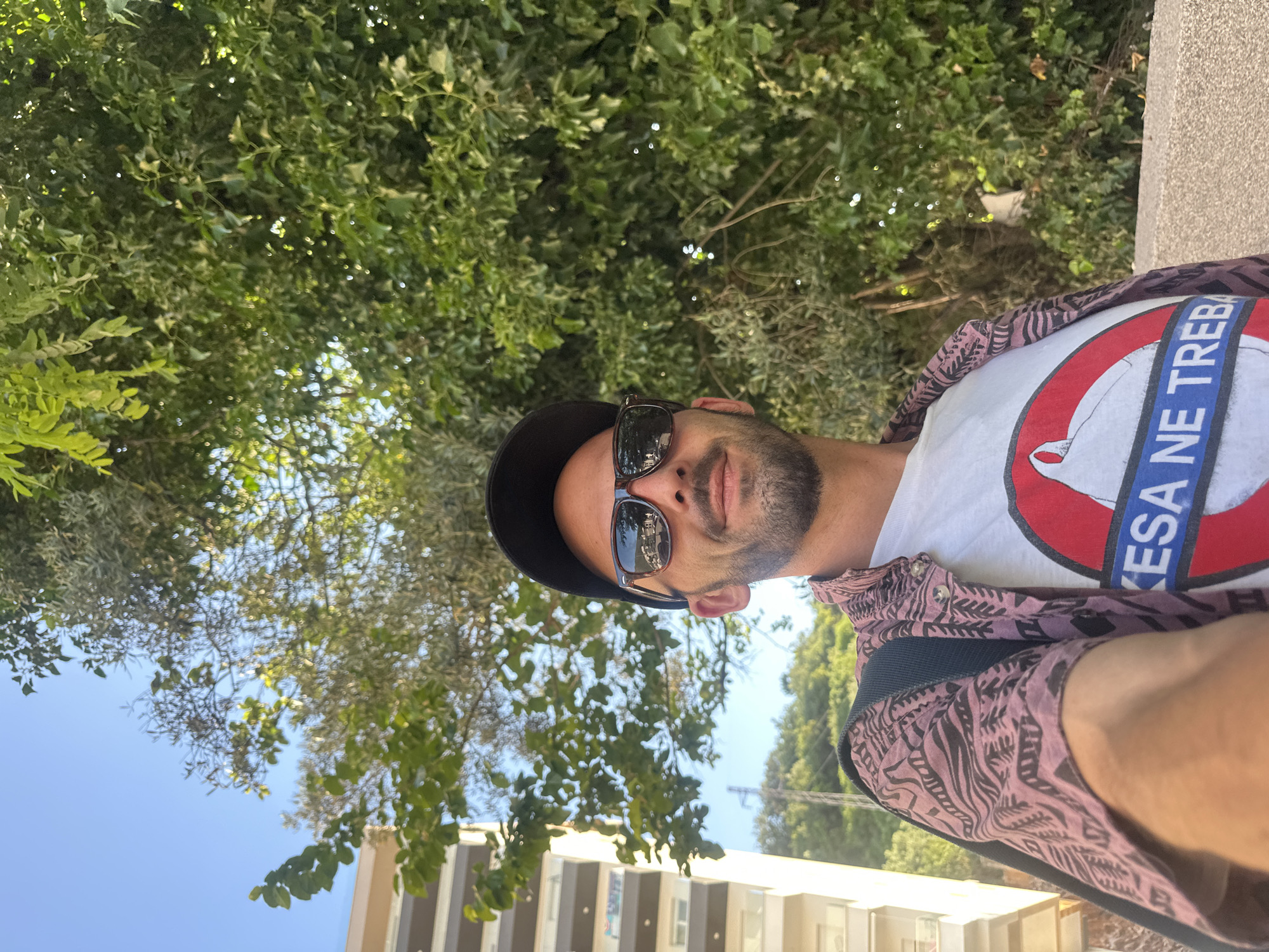 A man wearing a black baseball cap, sunglasses, a pink patterned open-necked shirt over a white t-shirt with a red and white circular logo that reads "KESA NE TREBA" and a black backpack is looking at the camera outdoors. He is in the city of Budva, Montenegro, with a tall, light-colored building to his left and lush green trees and foliage behind him. The sky is clear and blue.