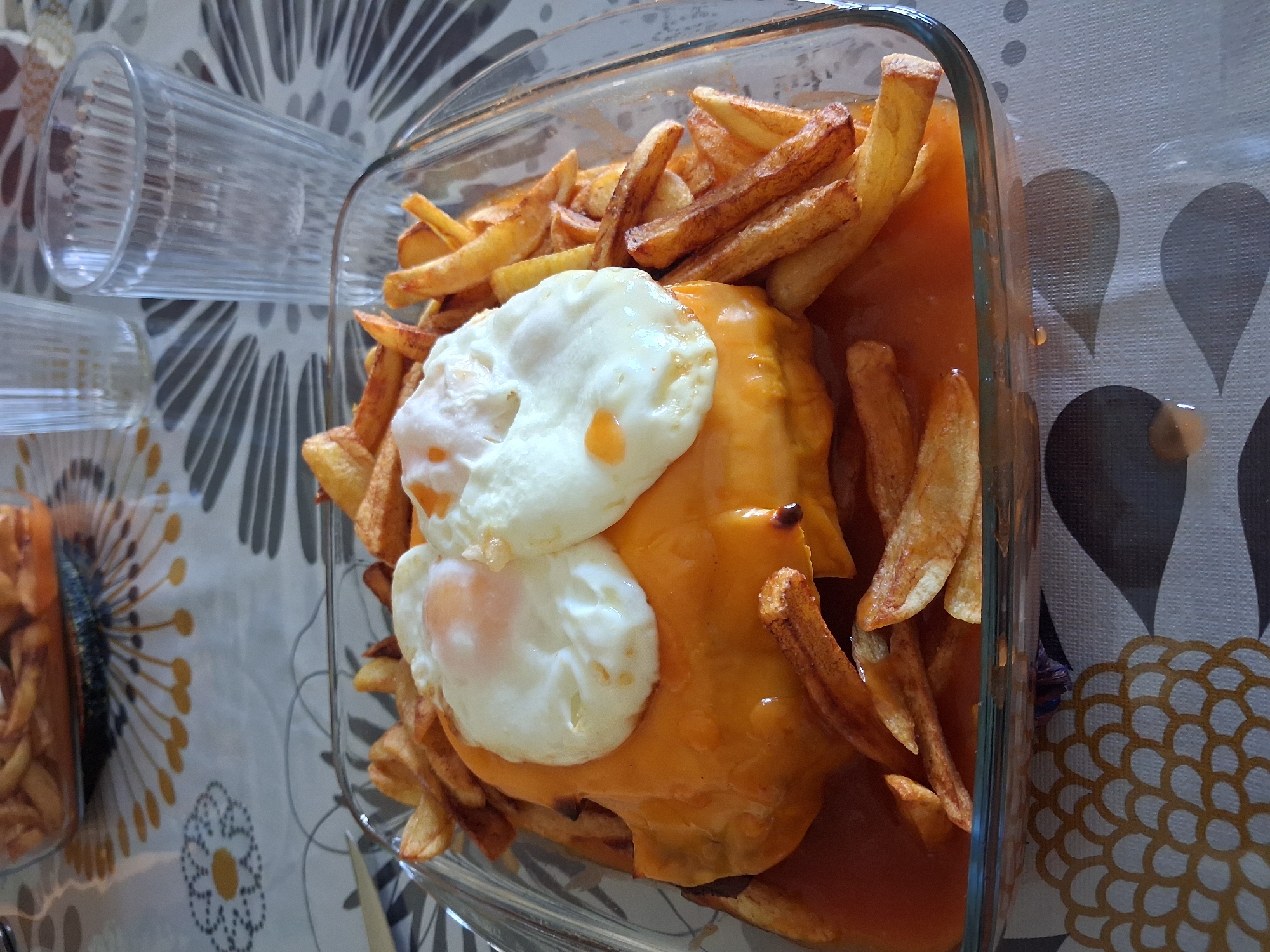 A Francesinha, a Portuguese sandwich, sits in a clear glass baking dish. Two fried eggs top the sandwich, which is covered in melted cheese. The sandwich is surrounded by french fries and a reddish-brown sauce. To the left of the dish are two clear drinking glasses, and to the bottom left is another container with french fries. The dish is placed on a tablecloth with a geometric pattern of gray, white, and yellow. The location is Figueira da Foz, Portugal.