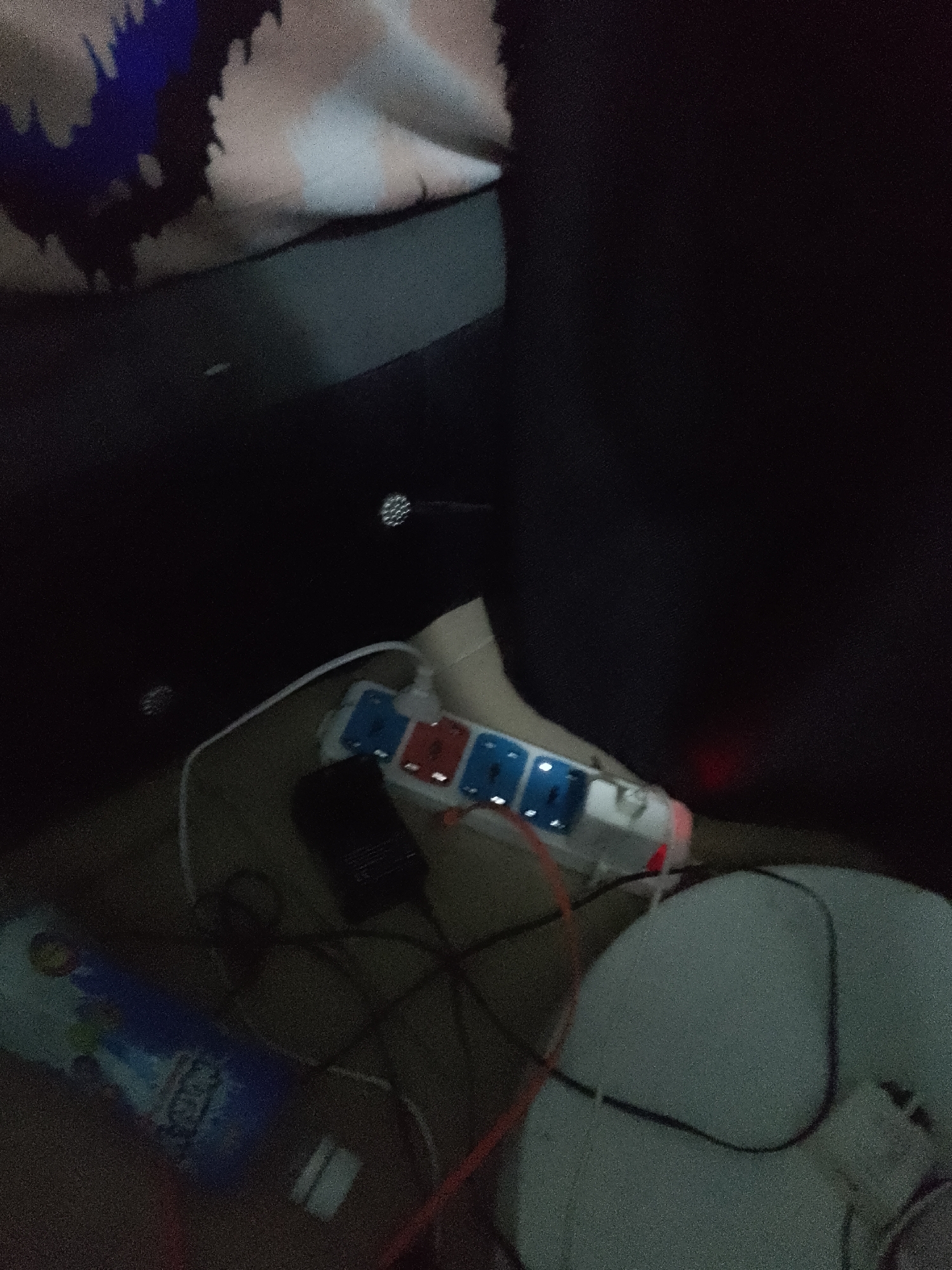 The image shows a close-up of a power strip with multiple outlets, some of which are occupied by plugs. The power strip is white with blue and red socket covers and is connected to various cords. There is also a black device, possibly a phone charger, plugged into one of the outlets. To the left of the power strip, a blue and white patterned bottle or container is visible, lying on its side. The background is dark and appears to be a fabric surface with a patterned design in shades of beige, white, and blue. A dark, solid object, possibly furniture, occupies the upper right portion of the frame. The scene is dimly lit.
