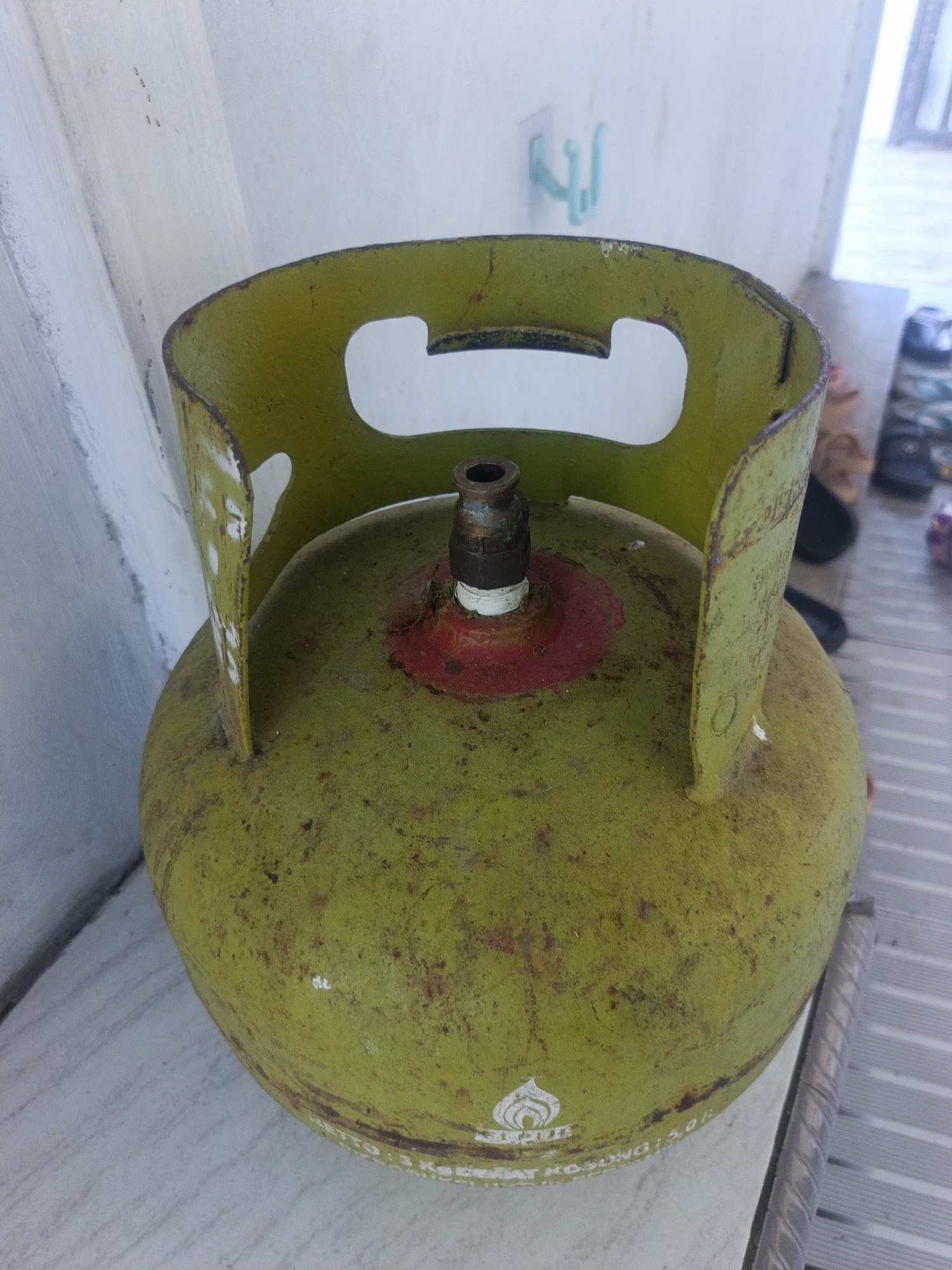 A lime green, cylindrical propane tank sits on a light-colored, horizontal surface. The tank has a rounded bottom and a slightly narrower top with a flared, circular collar that has two cutout handles. At the very top of the tank is a brass valve with a white ceramic piece. The tank has a weathered appearance with rust spots and scratches. There is white text printed around the bottom of the tank. Behind the tank, a white wall is visible with a light blue plastic hook. To the right of the tank, a metal grate or railing is visible, and further in the background, there are blurred shapes that appear to be shoes and possibly furniture. The scene is likely outdoors, possibly on a porch or patio, given the grate and the wall texture. The image is taken from a slightly elevated angle, looking down at the tank.