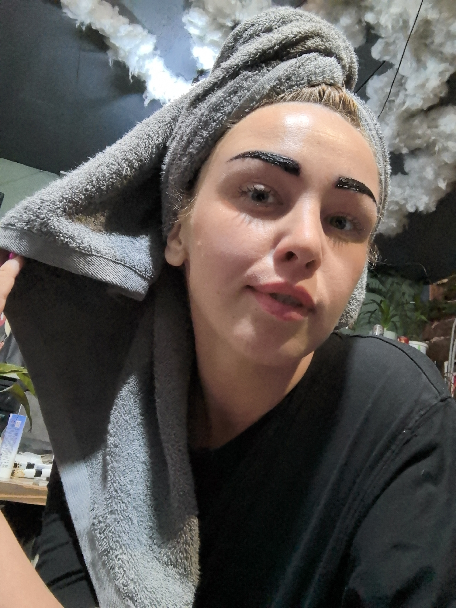 A woman is shown in a close-up selfie. She has a gray towel wrapped around her head. Her eyebrows have a thick, dark substance applied to them, presumably dye. She is wearing a black T-shirt. The background shows an indoor space with white, cloud-like decorations on the ceiling and some plants. There are also various objects on a shelf visible in the background.
The location is not in Noviy, Russia.