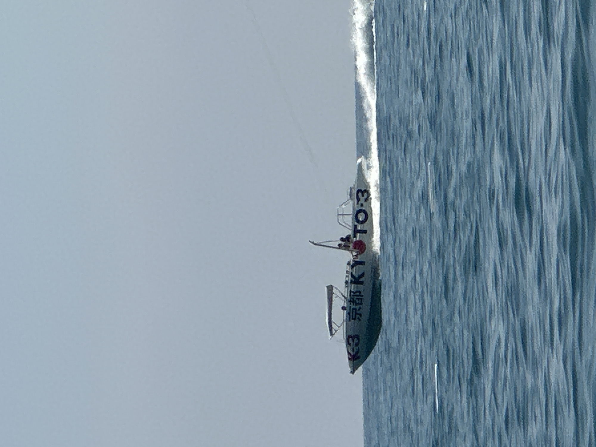 A speedboat with the markings "K-3 京都 KYOTO-3" is speeding across the water. The boat appears to be light gray with a dark stripe and has a canopy providing shade. There are at least two people visible on the boat, one standing near the stern and another seated. The boat is creating a significant wake, indicating it is moving at a high speed. The water is a deep blue, and the sky is a hazy light blue, suggesting a clear but possibly humid day. The scene appears to be an open body of water, likely a sea or a large lake. No specific landmarks identifiable to Boreti, Montenegro are visible.