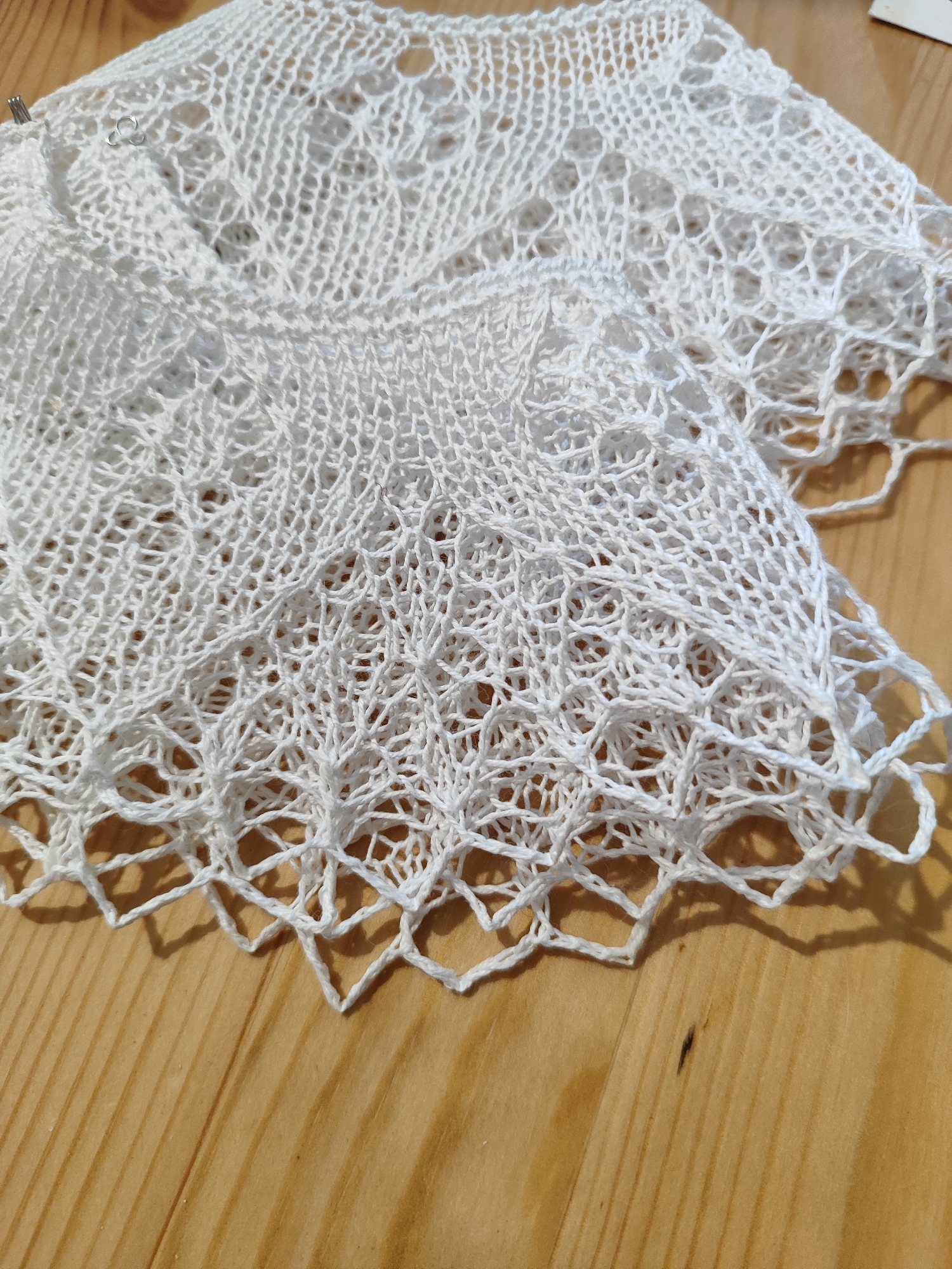 A close-up, eye-level, overhead shot shows a white, intricate, lace-like knitted fabric resting on a light-brown, wooden surface. The fabric is draped and folded, revealing complex stitch patterns and openwork details, with a decorative scalloped edge along the bottom. A few small metal stitch markers are visible near the top left of the fabric. The wooden surface exhibits a natural grain pattern with subtle variations in color and texture. The lighting casts soft shadows on the wooden surface, highlighting the texture of the fabric and the wood. There are no people or any other objects visible in the scene. The image does not provide any information or clues that would allow for the determination of its location within the city of Vilnius, Lithuania.