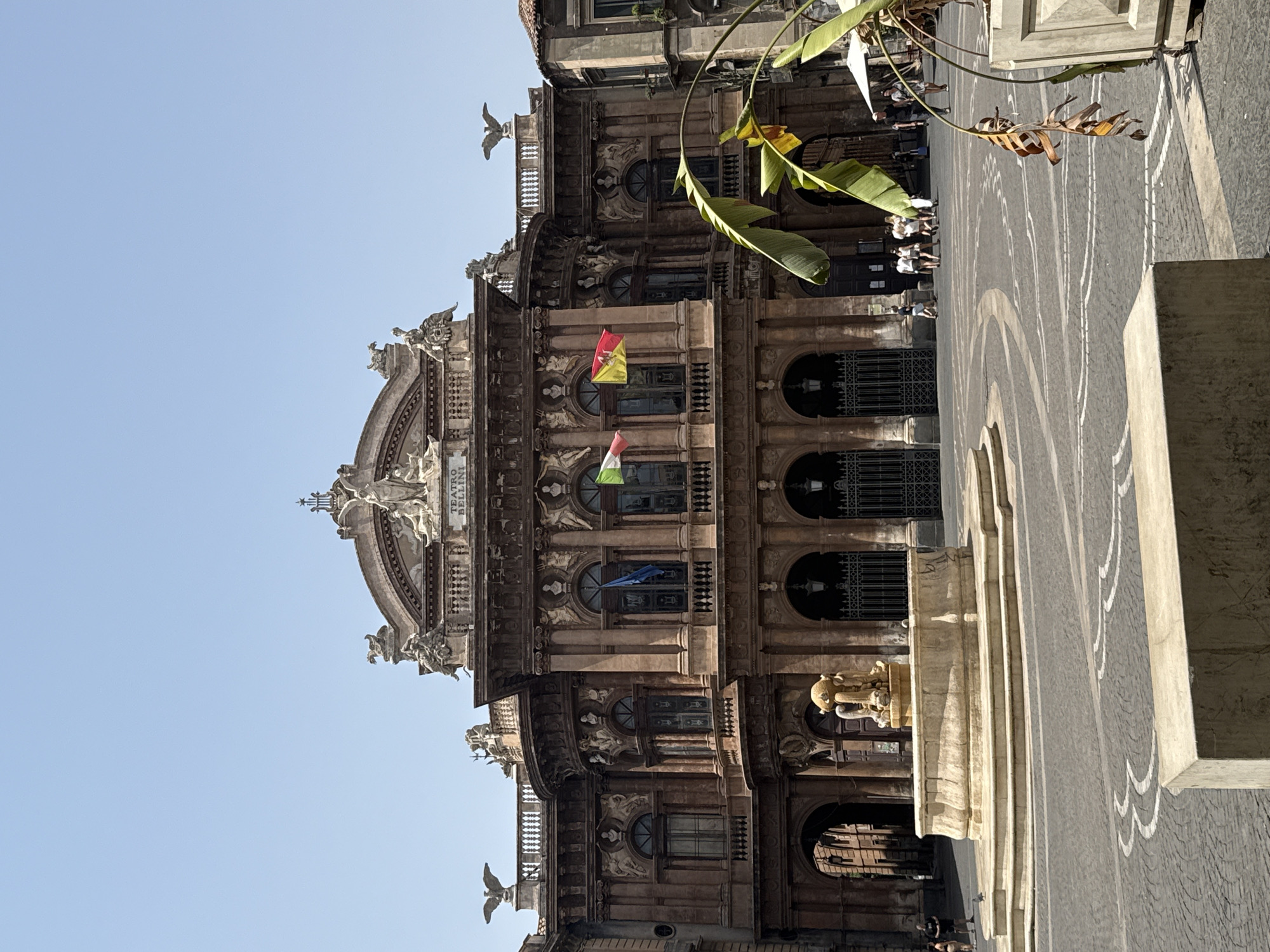 The image shows a wide-angle view of Piazza Teatro Massimo in Catania, Italy. In the center is the grand facade of the Teatro Massimo Bellini, an opera house with ornate architectural details, including statues and columns. Two flags, the Italian flag and the flag of Sicily, fly from the building. In the foreground, a multi-tiered stone fountain with a sculpture at its center is prominent. The plaza is paved with dark cobblestones and features decorative curved white lines. Several people are visible in the background, walking around the plaza and near the entrance of the theater. To the right, large green leaves of a tropical plant in a planter partially obscure the view of the opera house and the sky above is clear and blue.