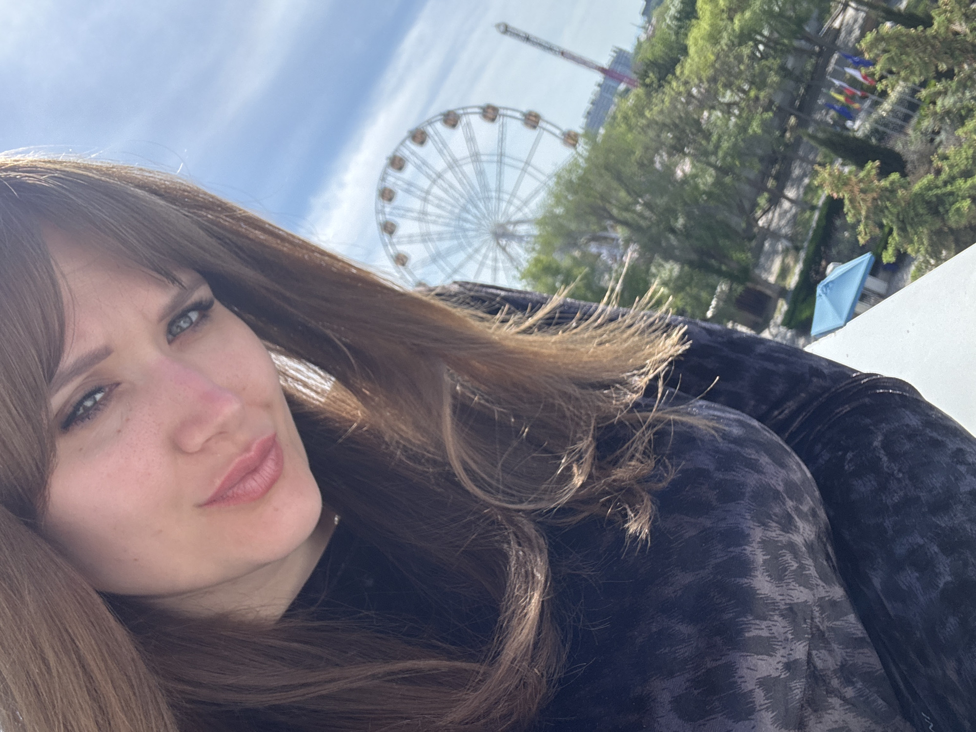 A selfie of a woman with long brown hair, fair skin, and freckles. She is wearing a dark patterned top. In the background is a Ferris wheel and a tall tower structure, along with trees and flags. The location appears to be a park or amusement area in the city of Yevpatoriya, Ukraine.