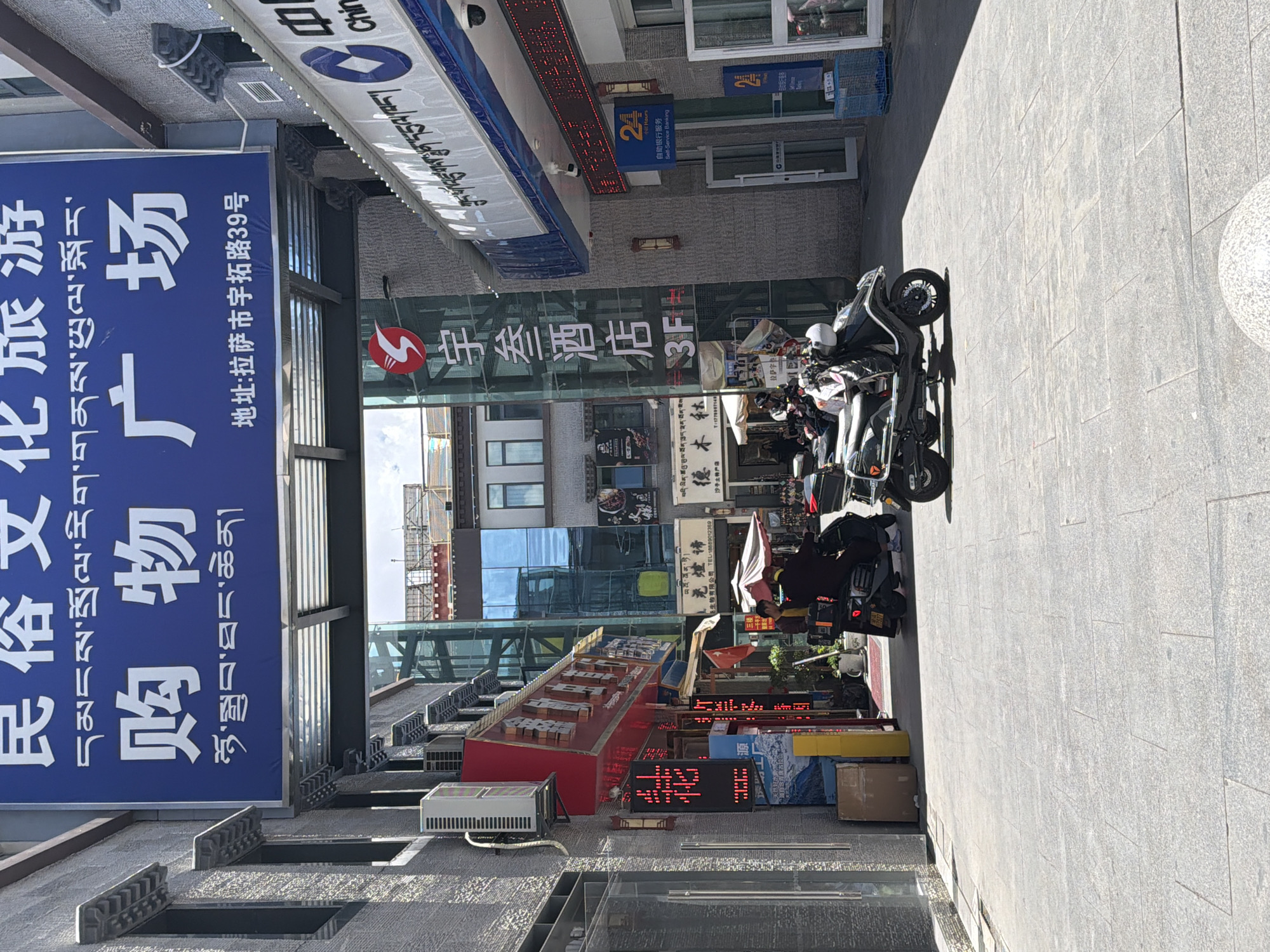 This daytime photograph captures a commercial street scene in Lhasa, China, specifically at No. 39 Yutuo Road, as indicated by visible signage. The perspective looks down a paved street lined with multi-story buildings on both sides. The bright sunlight casts sharp shadows, suggesting clear weather.
On the left, a large blue banner, featuring both Chinese and Tibetan script, advertises a "Folk Culture Tourism Shopping Plaza." Below it, an air conditioning unit is mounted on the building. The right side of the street features several parked motorbikes or scooters, some with white helmets. A few people are visible near these vehicles, with another person under an umbrella further down the street.
The buildings display various signs, including an overhead banner partially showing "Chin" (likely part of "China"). A prominent red LED sign above a banking facility indicates "24 Hours Self-Service Banking" in both English and Chinese. Another sign identifies "Yucan Hotel 3F." Other businesses advertised include "Demulin Sand Local Specialty Store," with names and phone numbers in both Chinese and Tibetan scripts. A red LED sign on the ground reads "源" (Source/Origin). The pervasive use of both Chinese and Tibetan languages serves as a strong location cue.