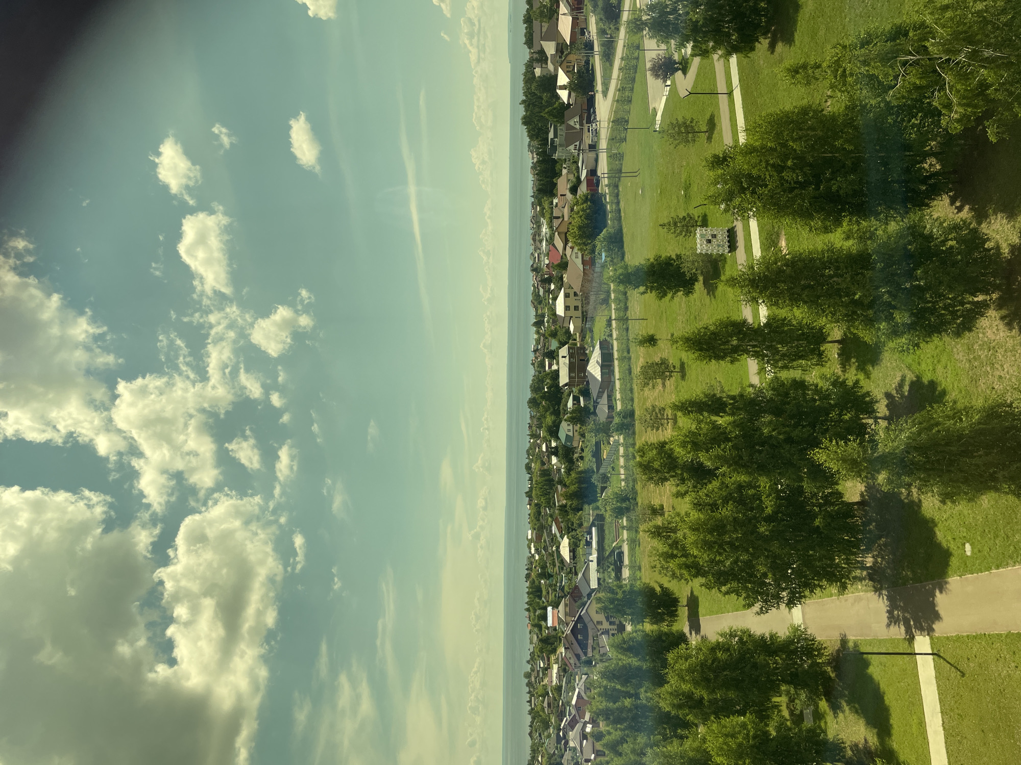This is a high-angle, medium-shot photograph of a residential area in Laishevo, Russia, on a partly cloudy day. The foreground features a park with manicured green lawns and scattered trees. A paved pathway winds through the park, with a lamppost visible along its edge. Beyond the park, a collection of houses with pitched roofs and varying architectural styles forms a village-like settlement. The houses are surrounded by greenery, and trees are interspersed throughout the neighborhood. In the distance, a wide expanse of water, likely the Kama River, stretches across the horizon, meeting the sky under a pale blue and grey cloudy atmosphere. The overall scene depicts a tranquil, suburban landscape.