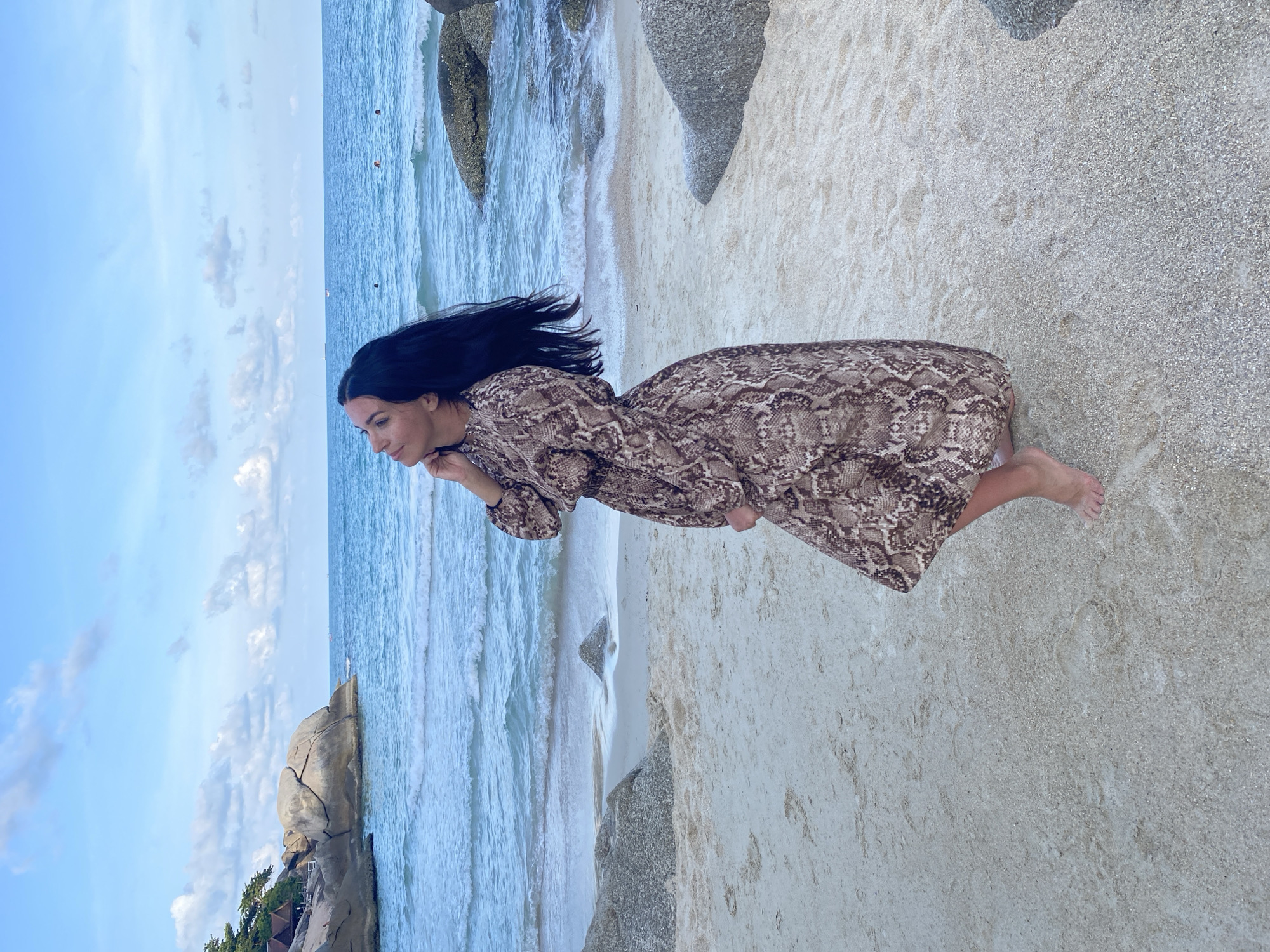 A woman with long black hair is standing on a sandy beach near the ocean, possibly in Ban Haad Rin, Thailand. She is wearing a long-sleeved brown dress with a snake-like pattern. Her hair is blowing in the wind. The ocean waves are visible in the background, along with some rocks and greenery. The sky is blue with some clouds.