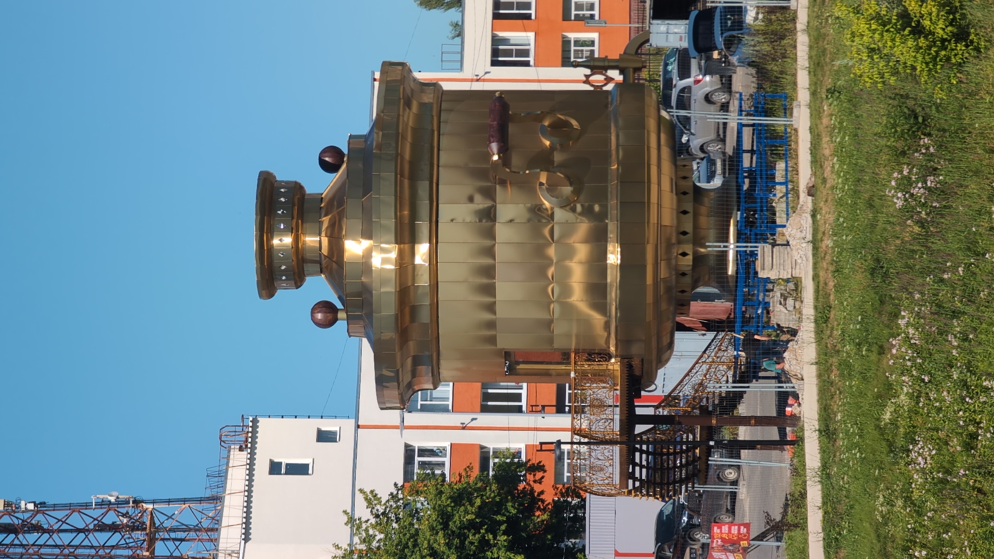 This image captures a large, ornate, brass-colored object resembling a samovar, placed outdoors in an urban or semi-urban setting. The object is positioned vertically, with its base towards the bottom of the frame and its top extending upwards. It appears to be a decorative installation or sculpture.
Surrounding the samovar, there are several buildings, including a white and orange multi-story building, and a building with white walls and orange accents. In the background, there are parked cars, including a silver SUV and a dark-colored van or SUV. A blue metal structure, possibly shelving or a temporary construction element, is also visible.
To the right of the samovar, there is a grassy area with some small pink wildflowers and green foliage. A pathway or sidewalk runs along the edge of the grass. In the foreground, there's a glimpse of what might be a construction site with metal scaffolding or framework.
The scene appears to be set during the daytime, under a clear blue sky. The lighting suggests it might be late afternoon or early evening, given the warm reflections on the brass. There are a few people visible in the lower part of the image, near the decorative metalwork in front of the samovar, one appearing to be wearing a teal shirt. One person seems to be working near the metal structure.
A few pieces of text are faintly visible on a red and yellow sign in the lower left, but the text is too blurry to be read. The overall impression is of a public space with a unique, oversized art piece. The location context points to Bor, Russia.