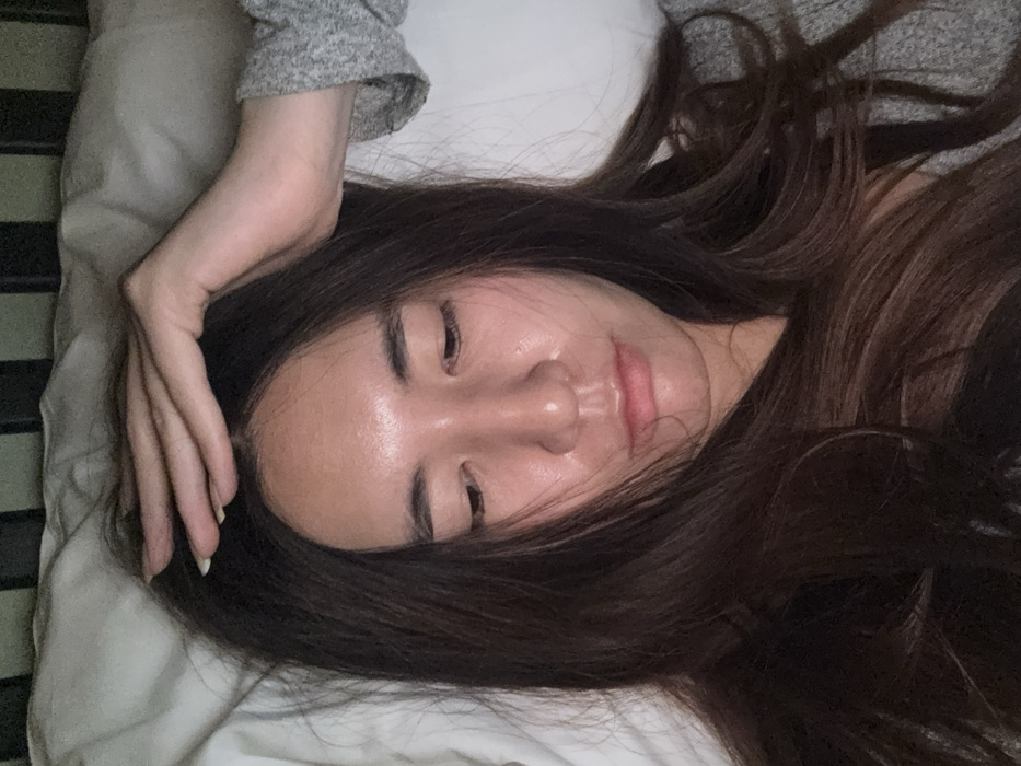 A young adult female is depicted in a close-up, overhead shot, lying on a bed indoors. She possesses long, dark brown hair, which frames her face and spreads across the white pillow. Her face is angled slightly downward and to the viewer's left, with eyes cast low or partially closed, and a neutral, slightly pensive expression. Her skin appears luminous. Her right arm is raised, with her hand resting on the top of her head and forehead, fingers subtly splayed. She wears a light gray, textured long-sleeved garment on her visible arm. The bedding consists of a white pillow and sheets. A dark, indistinct horizontal structure, possibly a bed frame or headboard, is visible above her head against the light background. The lighting is dim, suggesting an indoor setting. The image does not contain any visual information to determine a specific location within the city of Le Havre, France, or any other city.