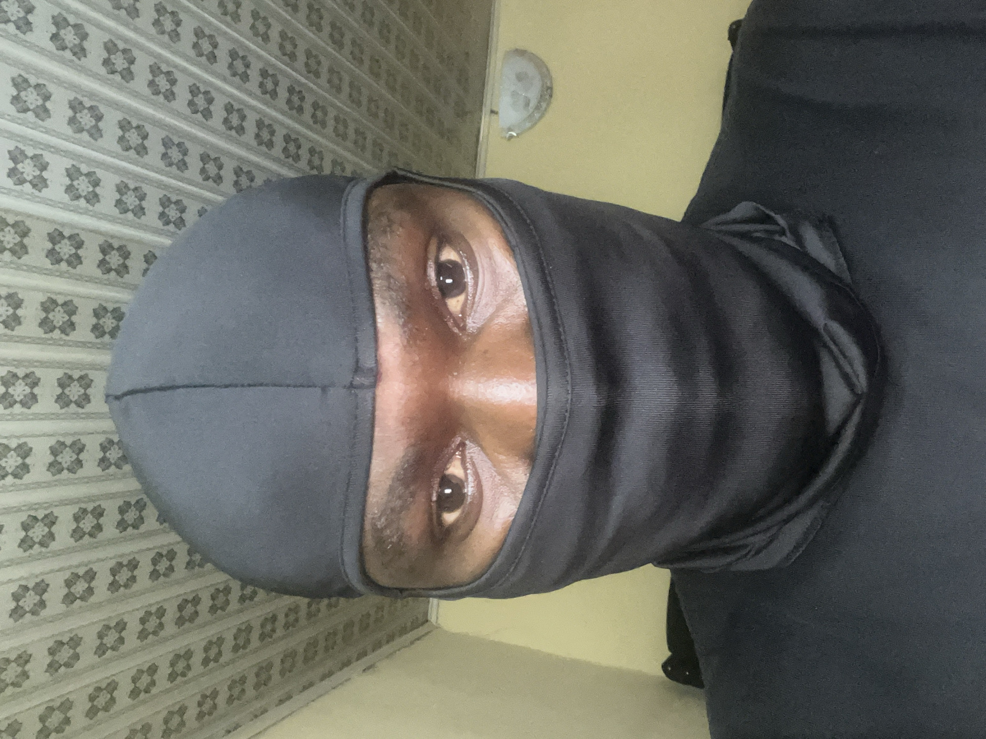 A person wearing a black balaclava is shown from the shoulders up, indoors. The background includes a floral patterned wall, part of an electric light fixture, and a plain painted wall. There is no information to ascertain if the location is in Jos, Nigeria.
